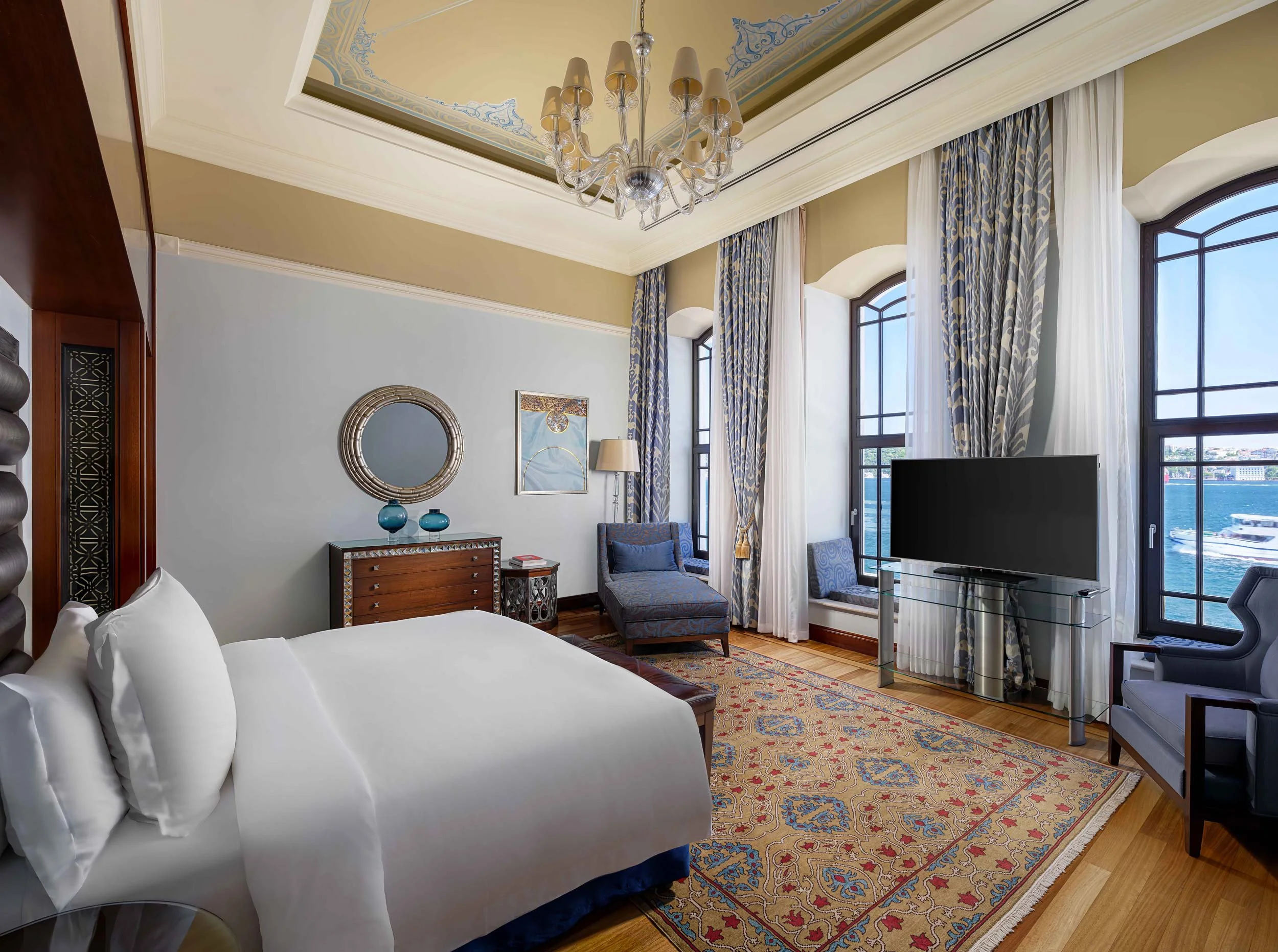 Luxurious hotel room with a large bed, a chandelier, a flat-screen TV, large windows overlooking water, and elegant furniture and curtains.