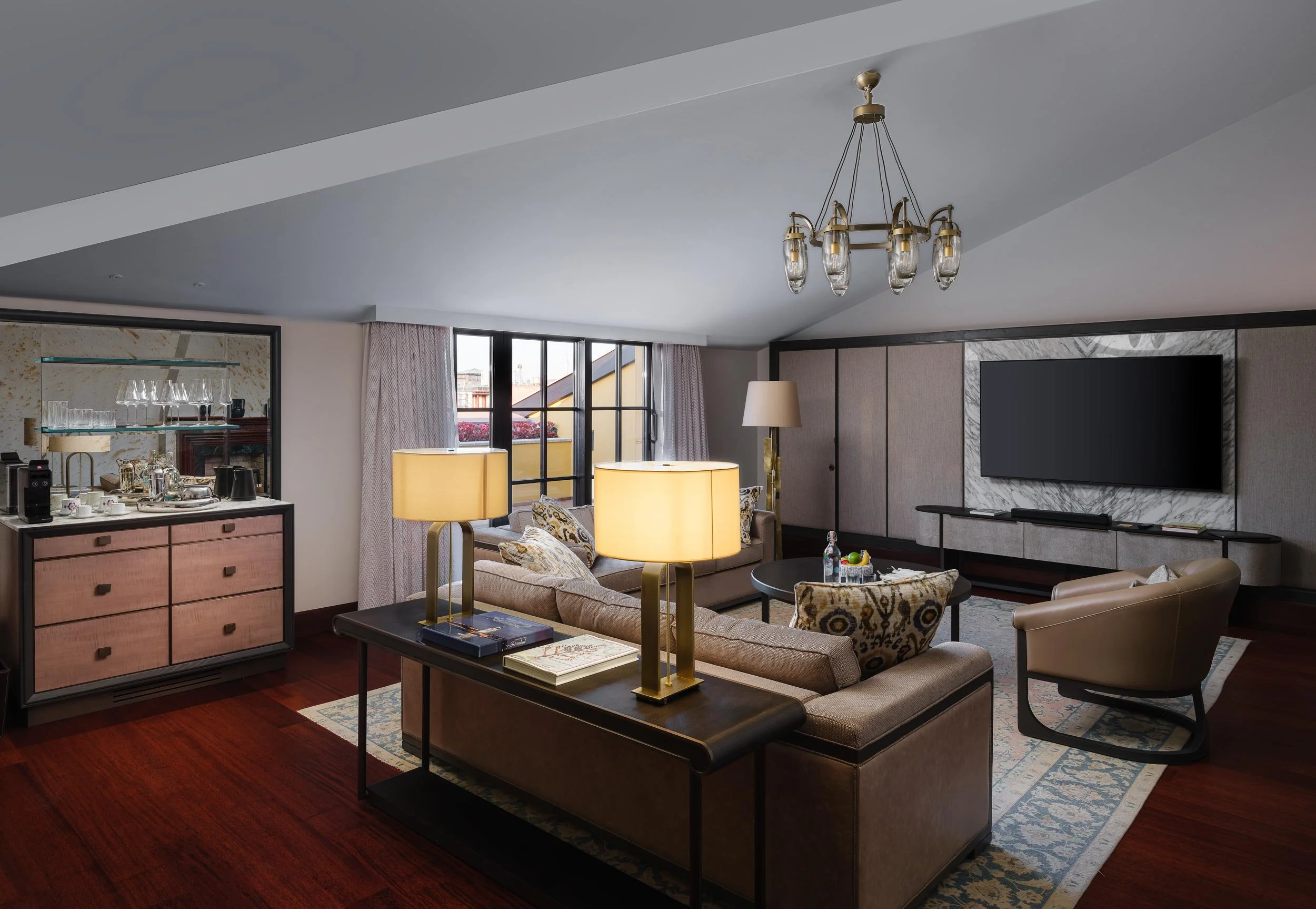 Elegant living room with beige sofas, a dark wooden coffee table, a large flat-screen TV mounted on a marble wall, and a chandelier hanging from the ceiling. There are table lamps on side tables, and large windows with curtains providing natural ligh