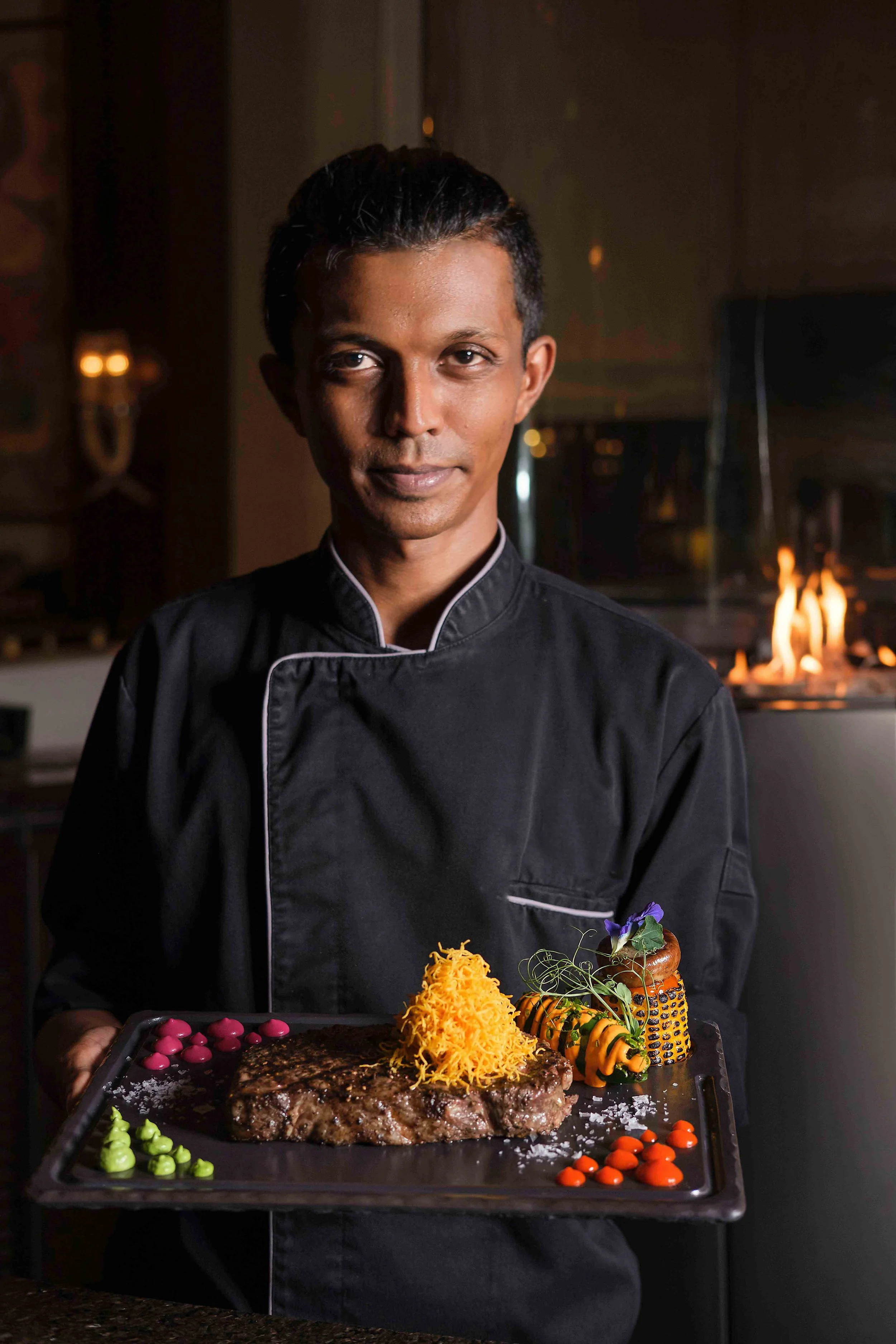 Chef holding a gourmet steak meal on a black tray, garnished with colorful sauces and vegetables.