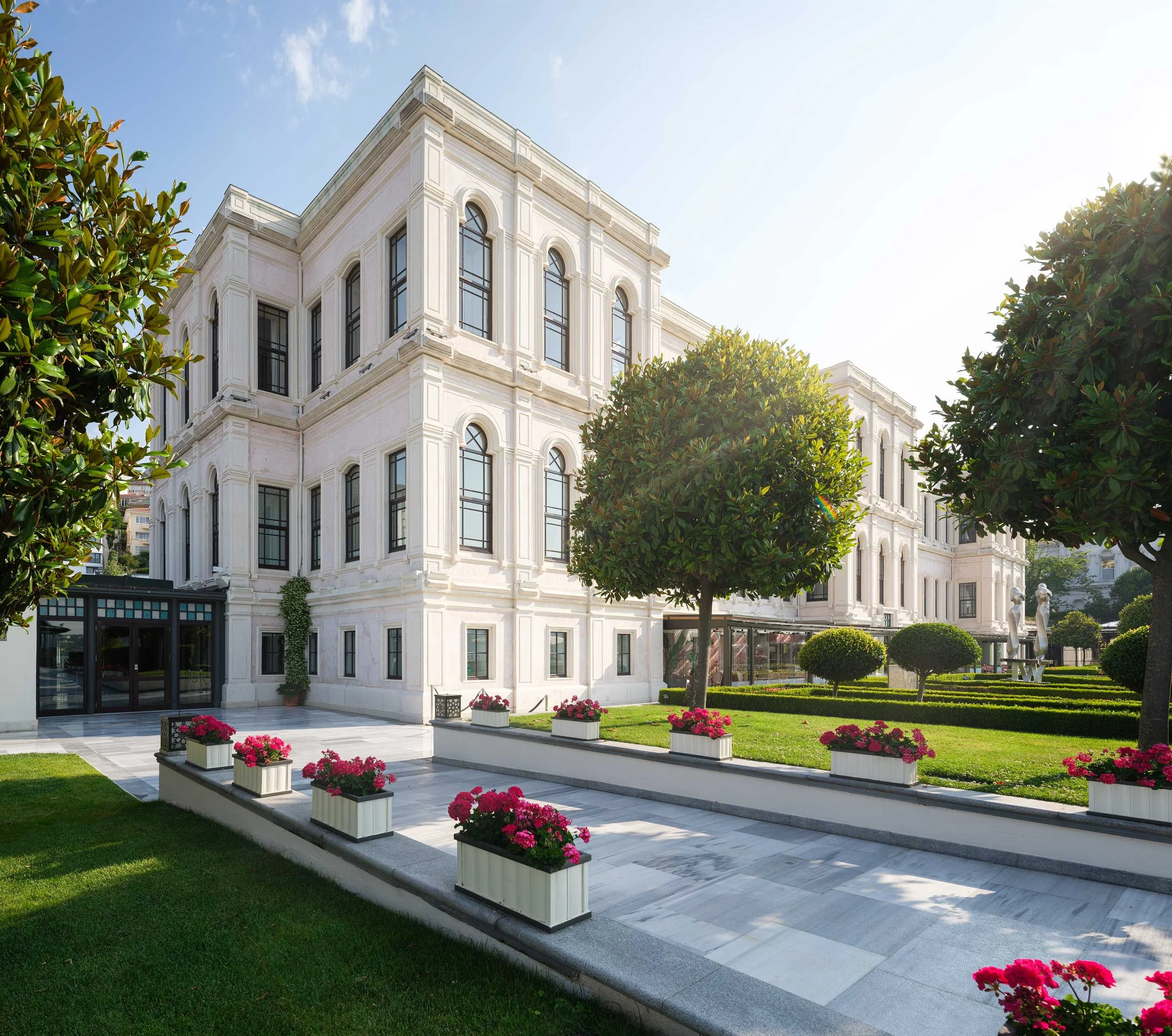 A large, white, three-story building with arched windows and decorative architectural details, surrounded by a landscaped garden with trimmed bushes and pink flowers, on a sunny day.