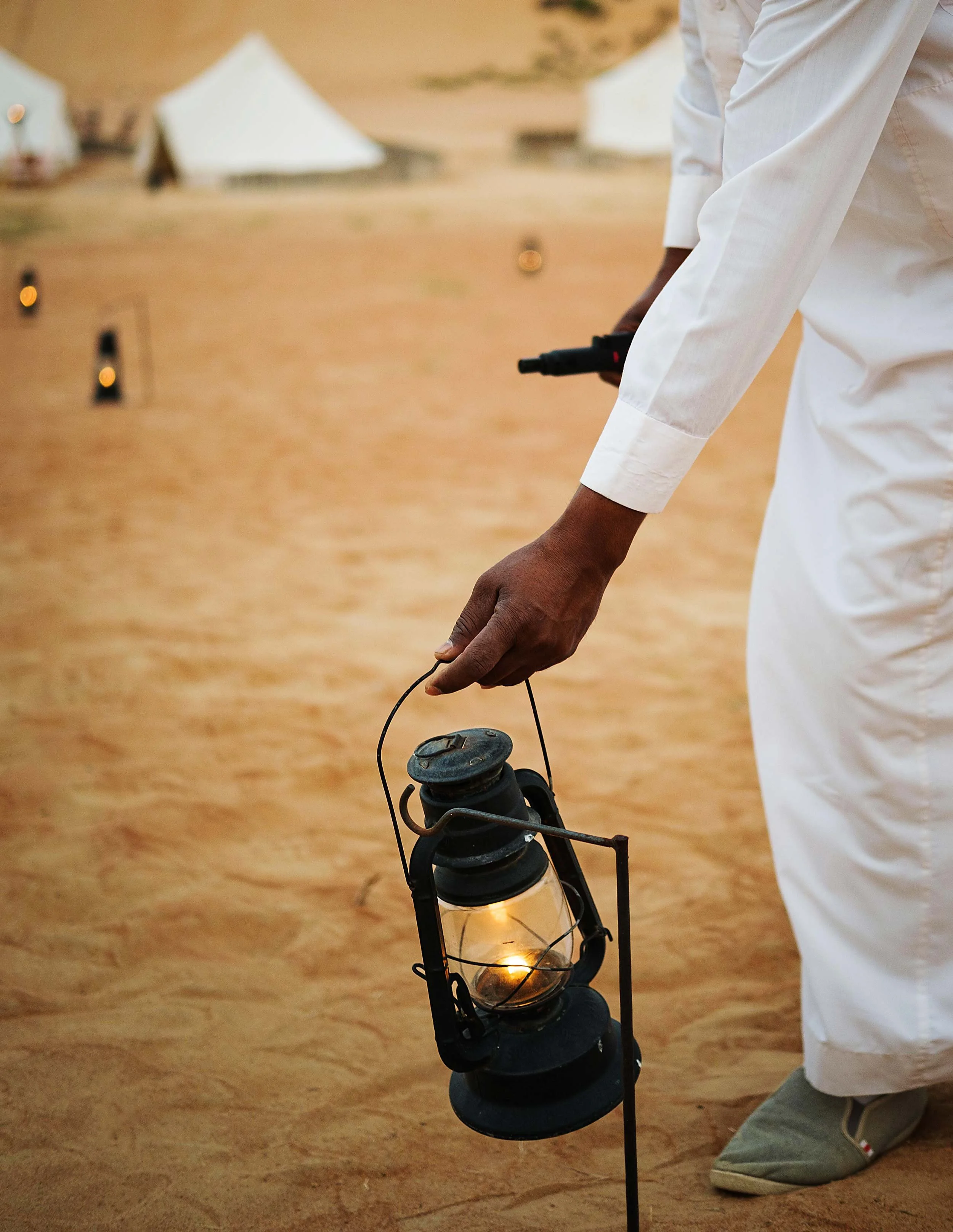 Person holding an old-fashioned oil lantern with a lit candle inside, standing on sandy ground with tents in the background.
