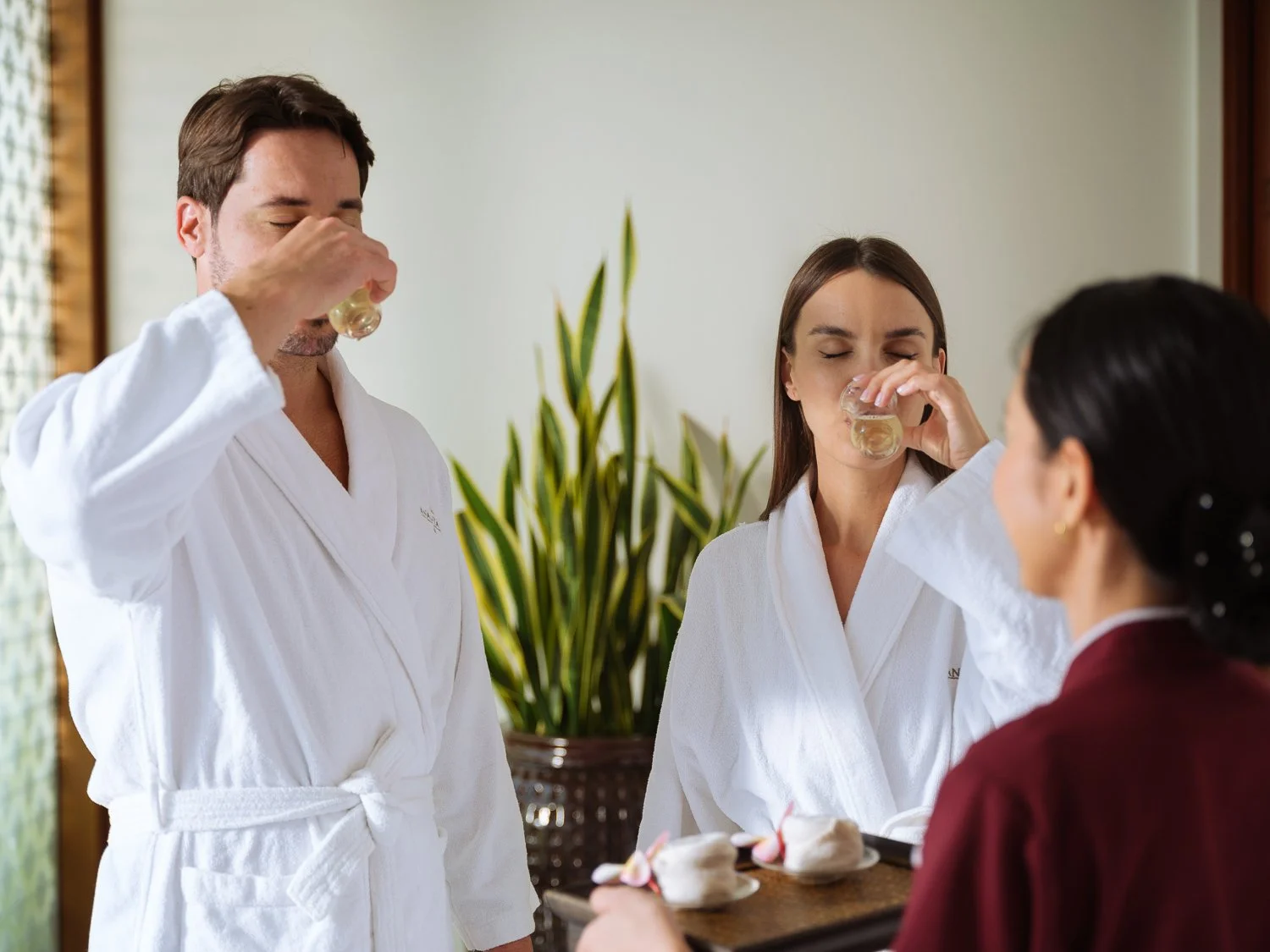 Three people in a spa setting; a man and a woman in white robes drinking from small glasses, served by an attendant holding a tray with towels and flowers.