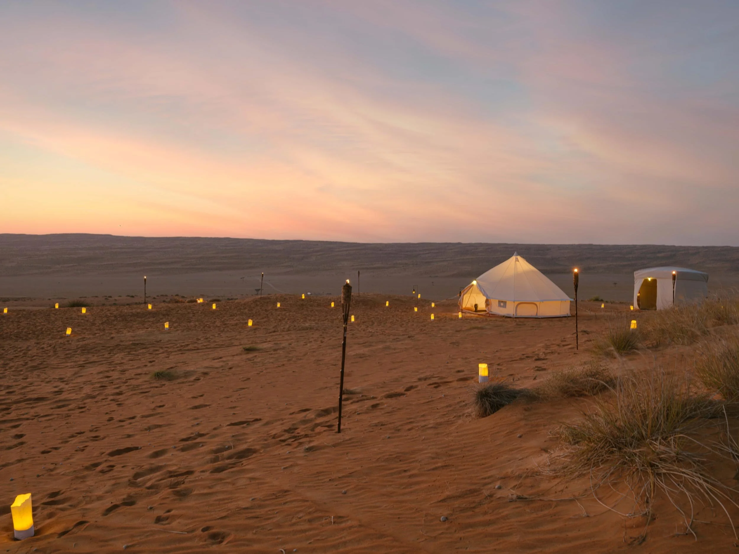 Desert camp with two white tents, illuminated lanterns scattered on the sand, during sunset or sunrise, with a flat horizon and a pastel sky in Bidiyah.