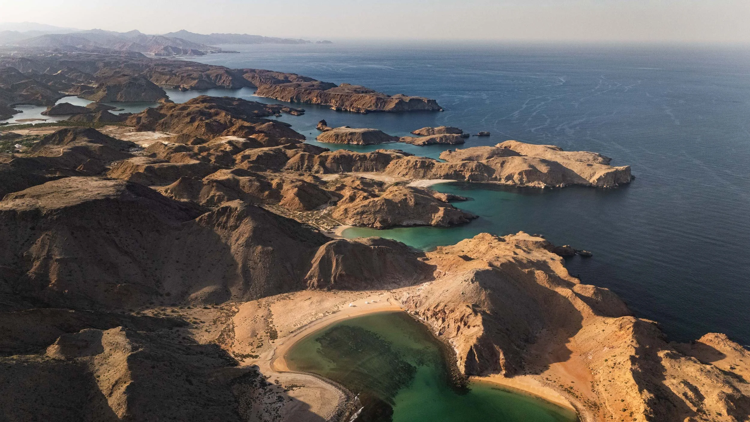 Aerial view of rugged brown hills and cliffs near the coastline with small bays and greenish water, possibly Lake Mead, with distant mountains and the open ocean in the background in bandar al khiran - Oman.