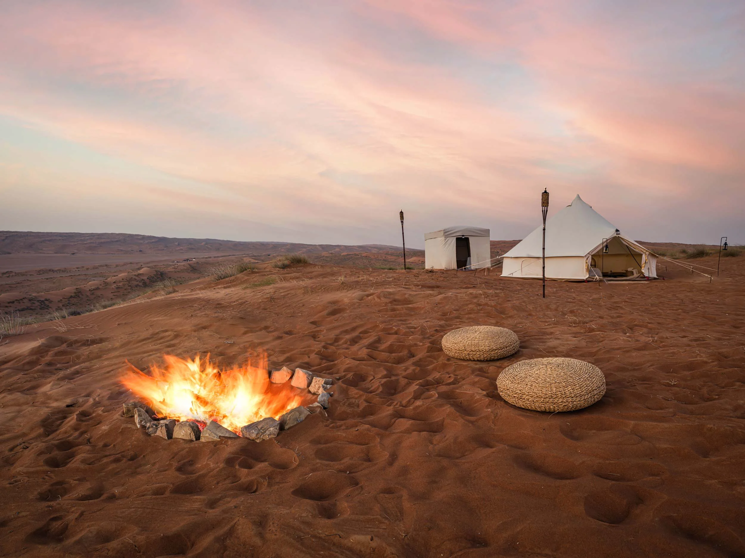 A desert campsite at dusk with a campfire surrounded by rocks, two woven ottomans, and white tents, with a sunset sky overhead.