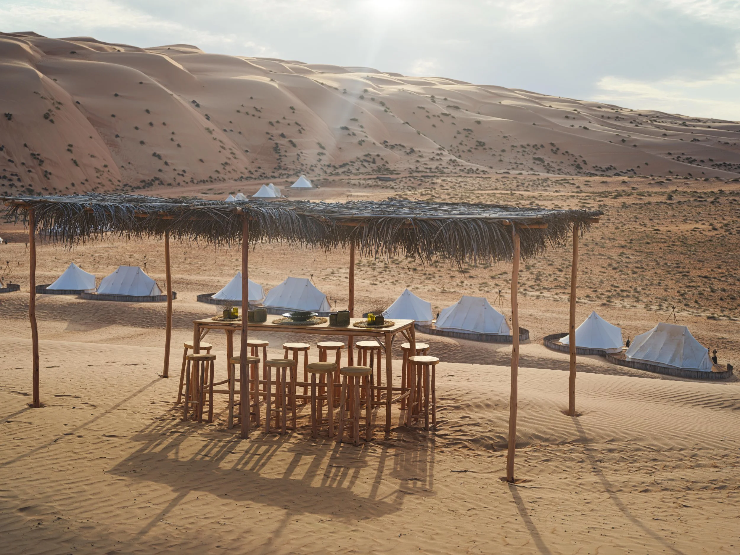 Desert camp with wooden stools and table under straw canopy, surrounded by white canvas tents on sandy terrain.