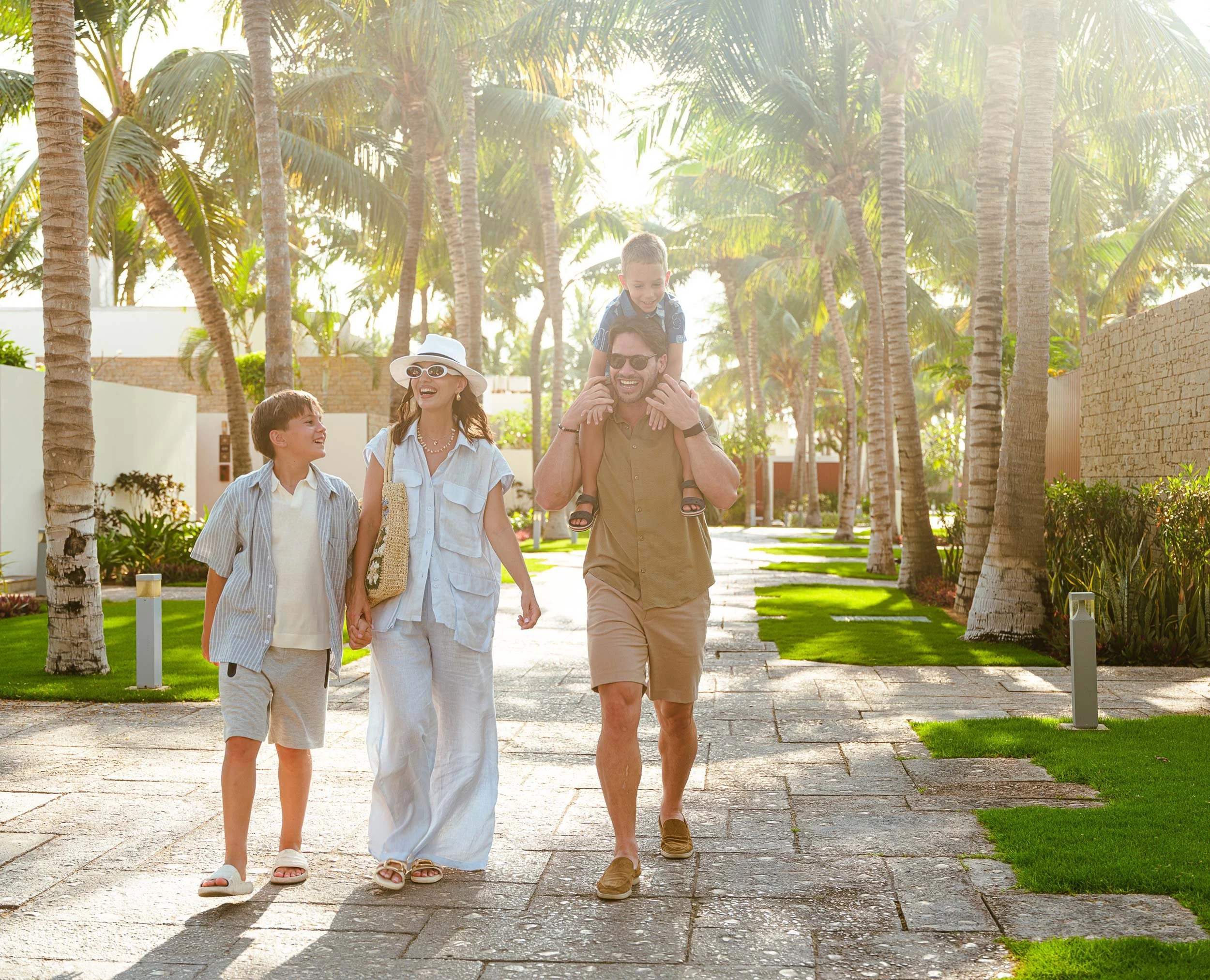 A family of four walking along a sunny pathway lined with palm trees, with the father carrying a young boy on his shoulders, the mother and an older boy walking beside them, all smiling and dressed in casual summer clothes.