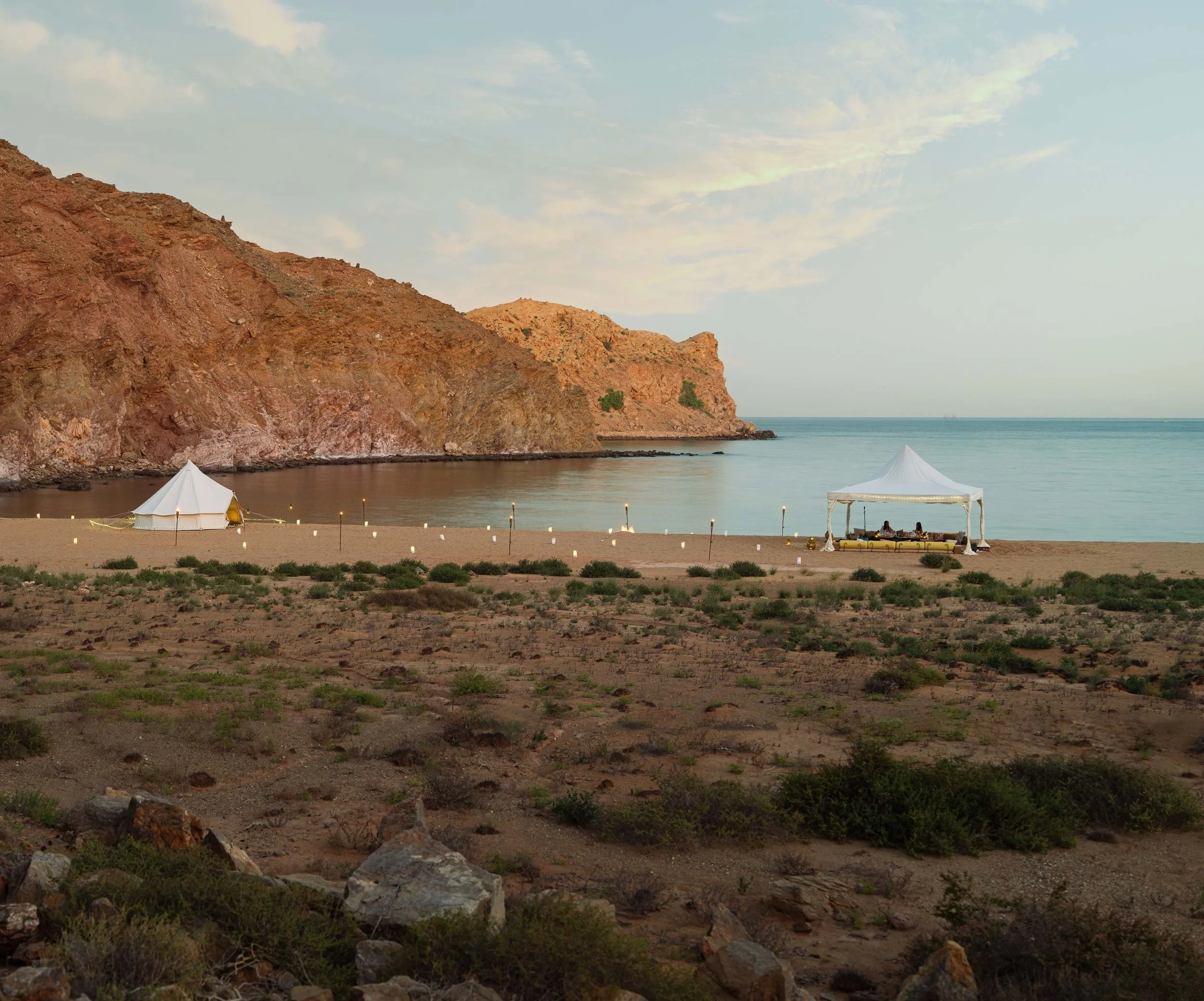 A beach scene with a white tent and a white canopy set up on the sand near the water, with mountains in the background and cloudy sky overhead in Bandar al Khiran - Oman.