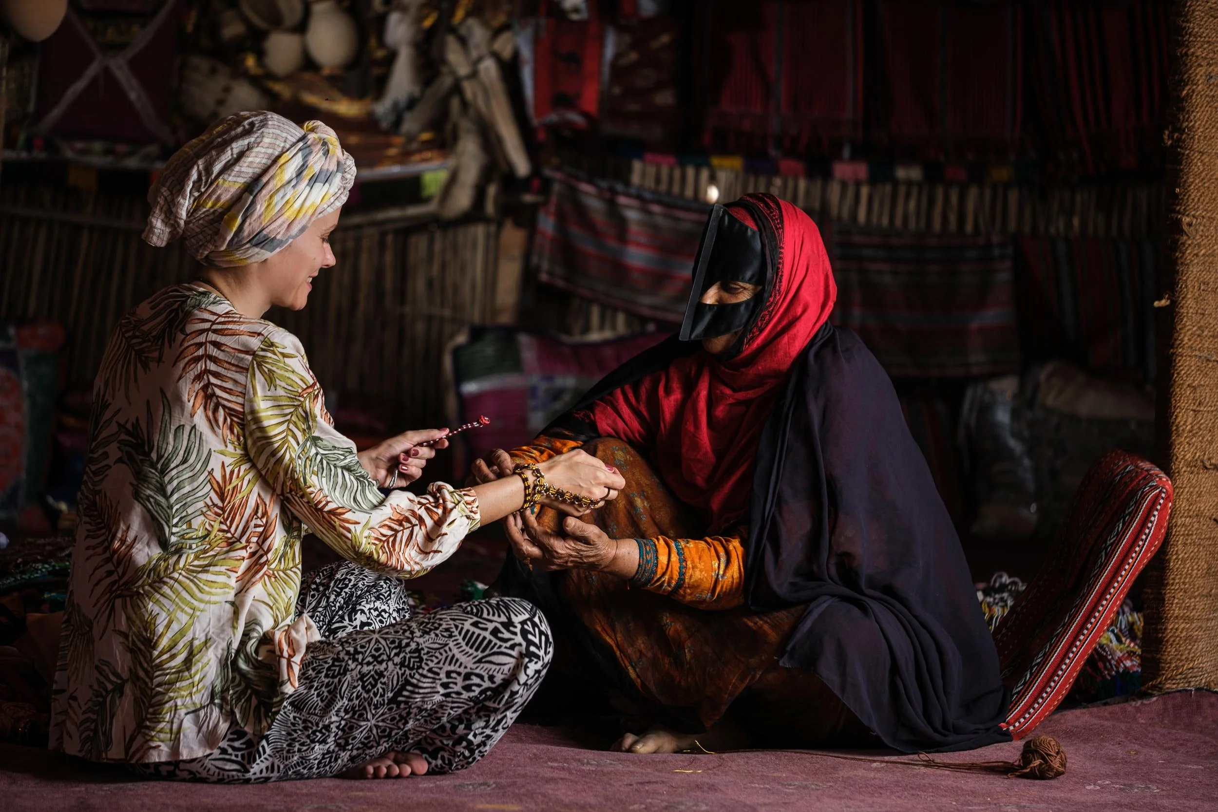 A woman in colorful traditional clothing and headscarf is sitting on the floor and offering a bracelet to a woman in black and red attire with a face covering, sitting on a cushion amidst a background of woven textiles.