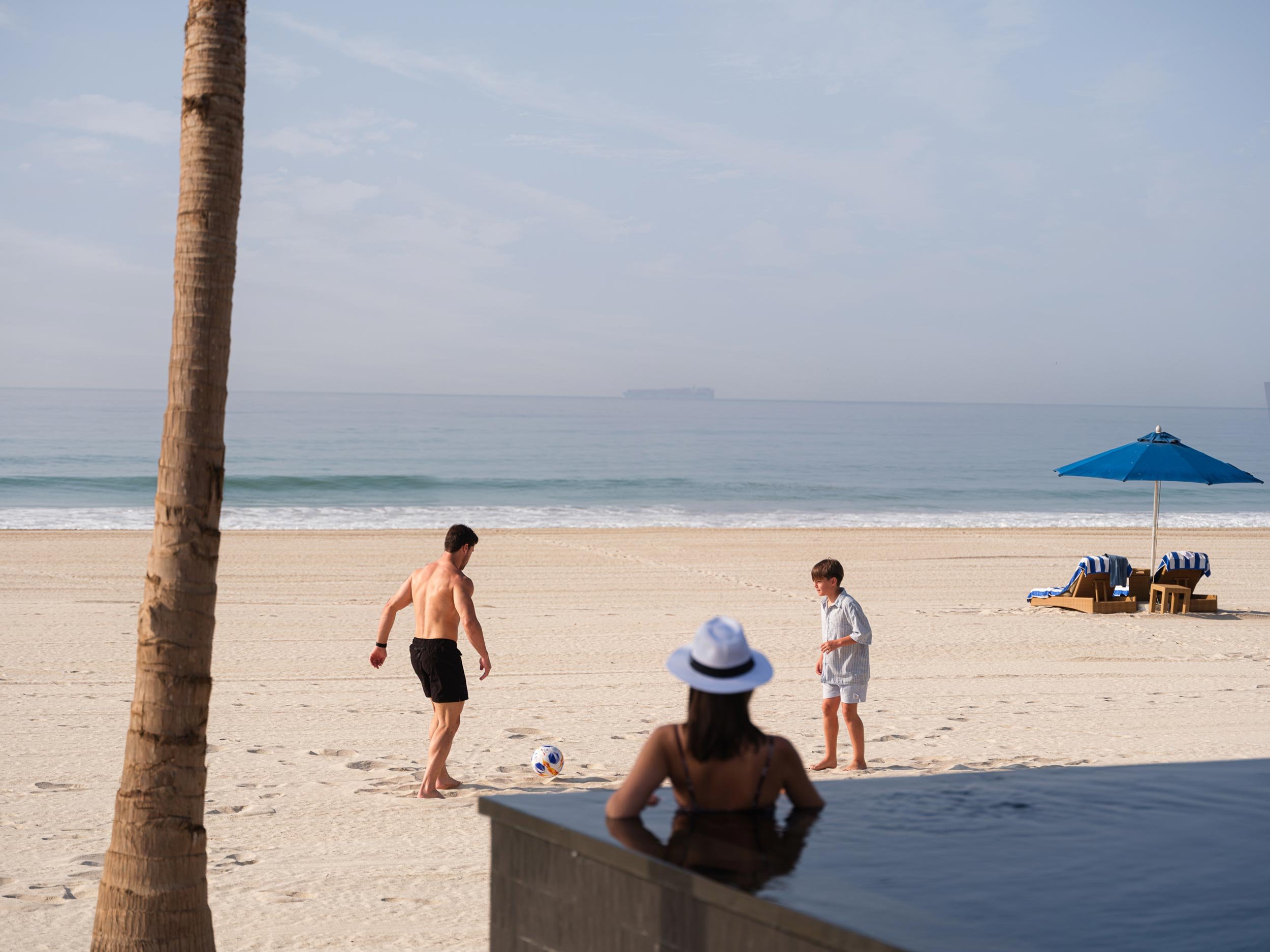 People playing soccer on the beach near the ocean, with beach chairs and an umbrella in the background.