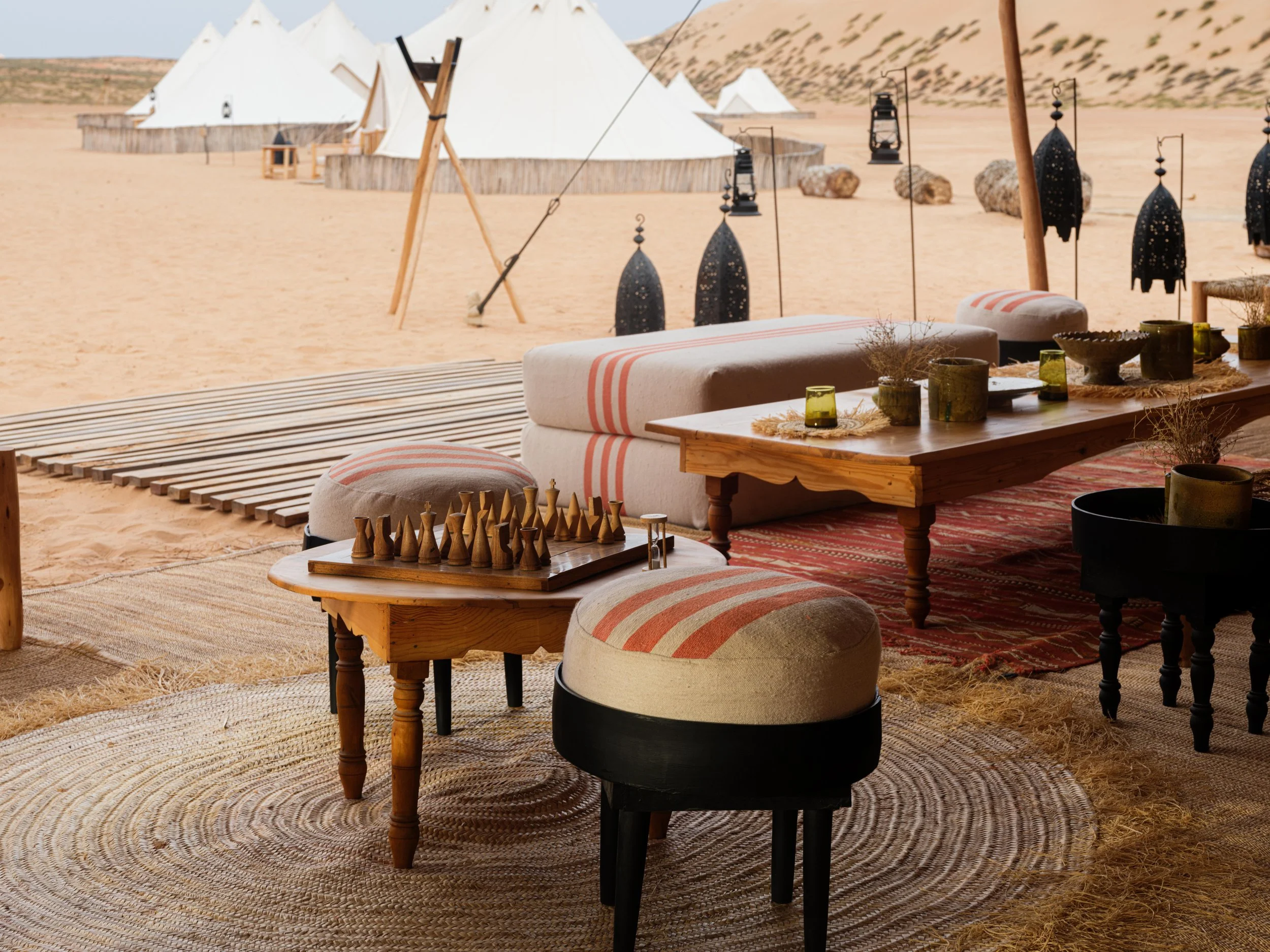 Desert camp setup with white tents and seating area, featuring striped cushions, a chessboard, and decorative lanterns on a sandy base.