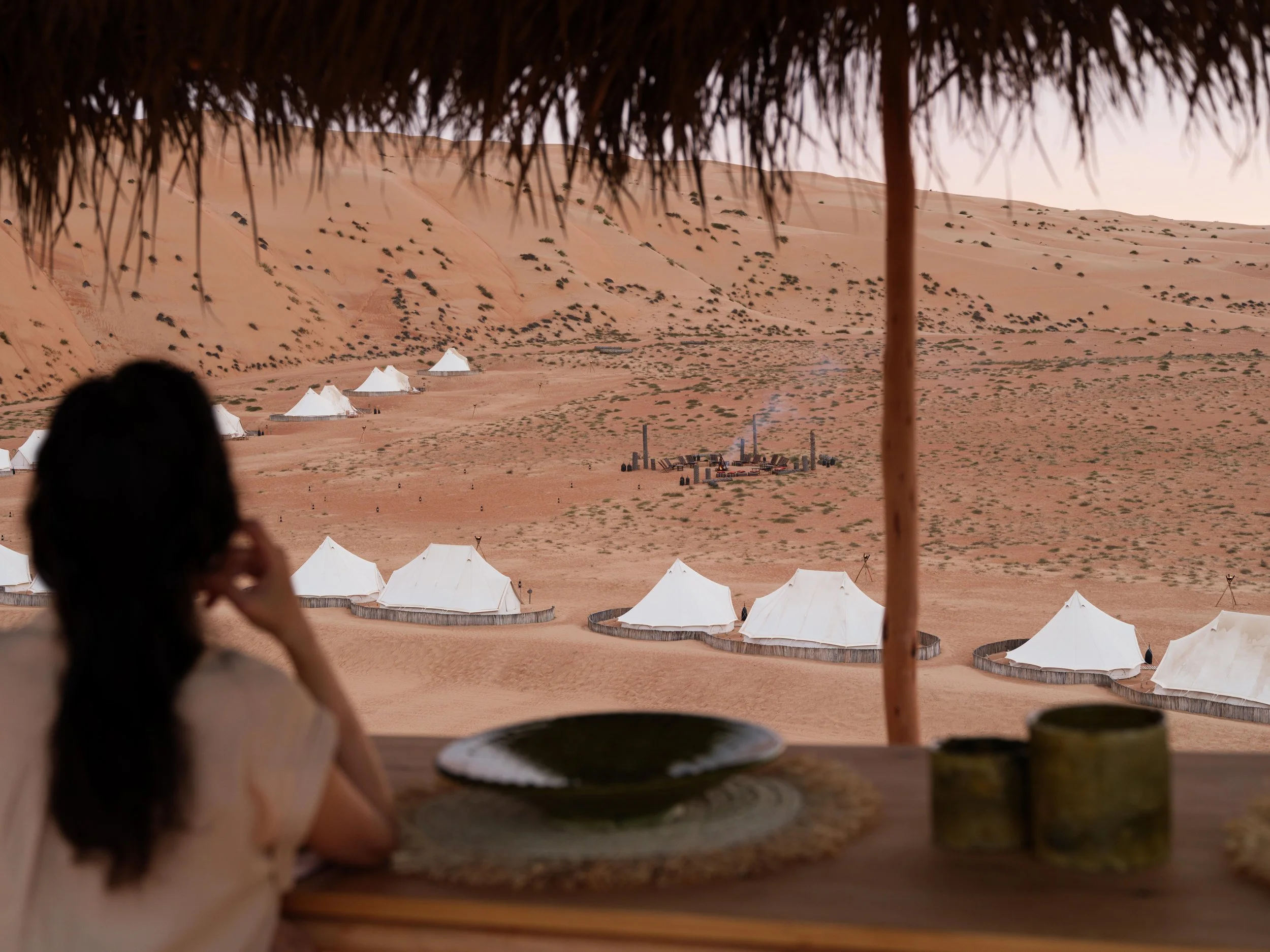 Desert glamping site with canvas tents and scattered greenery on sand dunes, with a person in the foreground viewed from a shaded vantage point.
