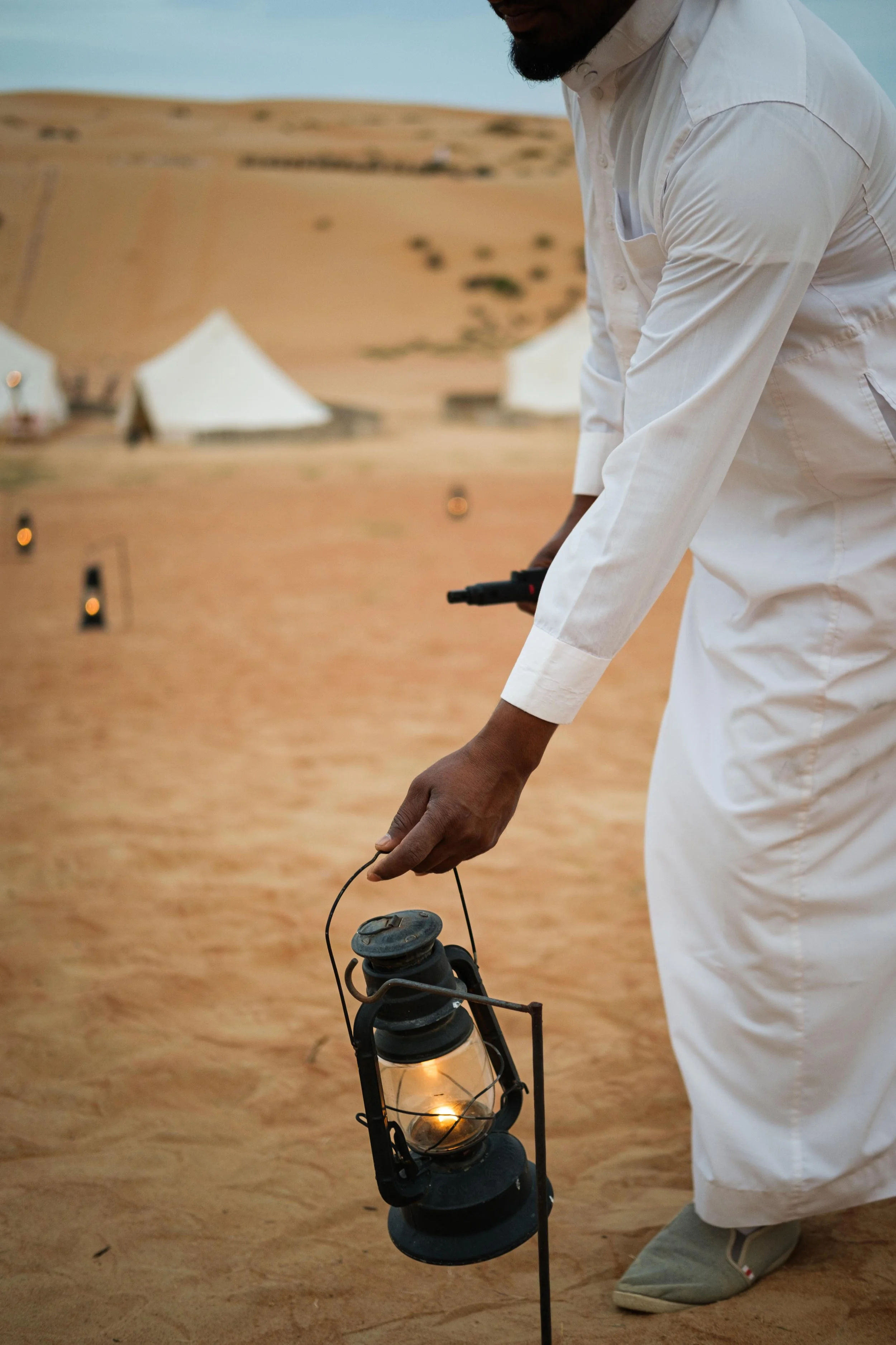 Man in white traditional attire placing a lit lantern on sandy ground with tents and desert in background.