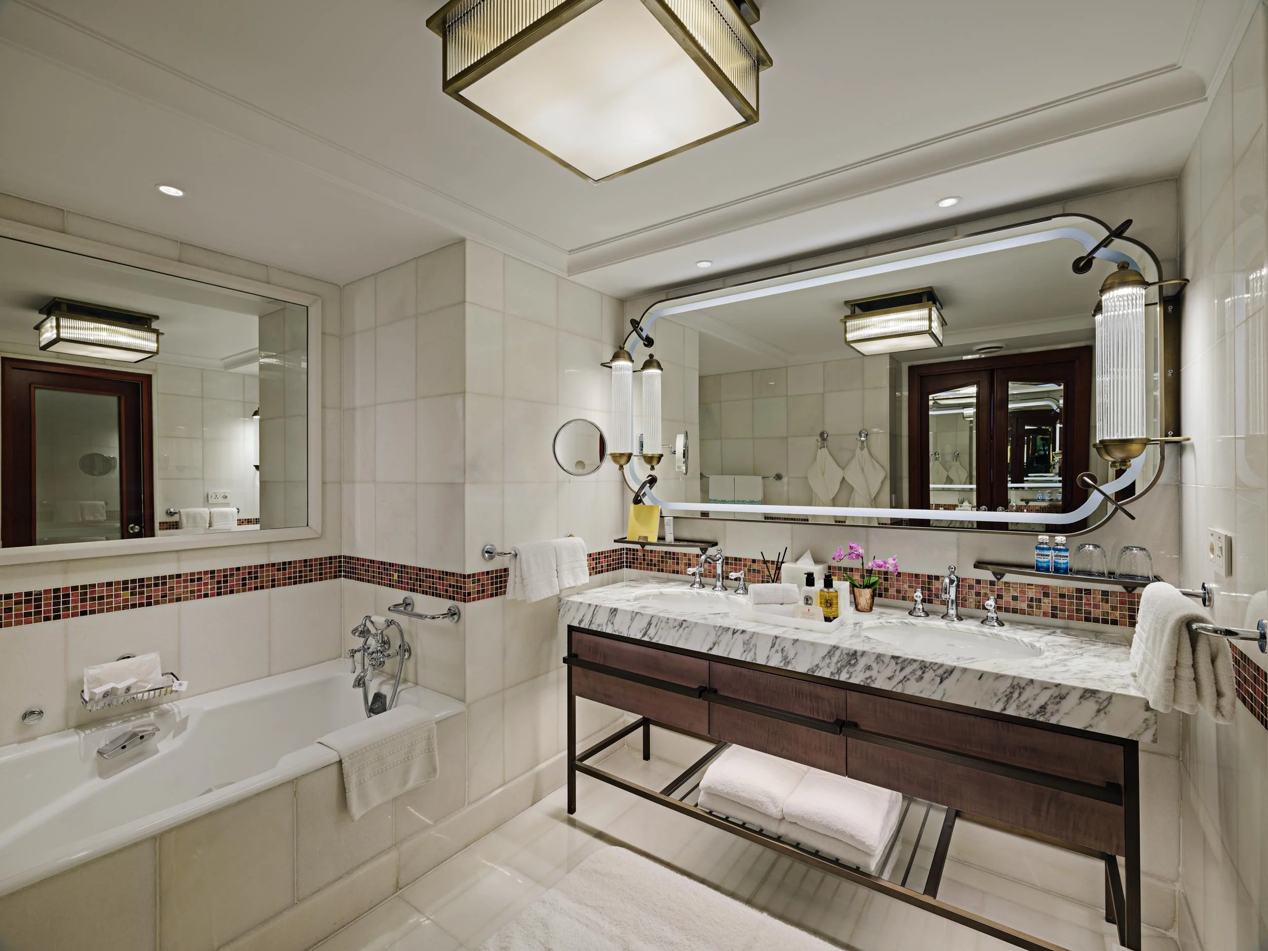A luxurious bathroom with a large marble double sink vanity, decorated with pink orchids, toiletries, and towels. Features a large mirror, wall-mounted lighting, a bathtub with a towel, and a mirror reflecting the room.