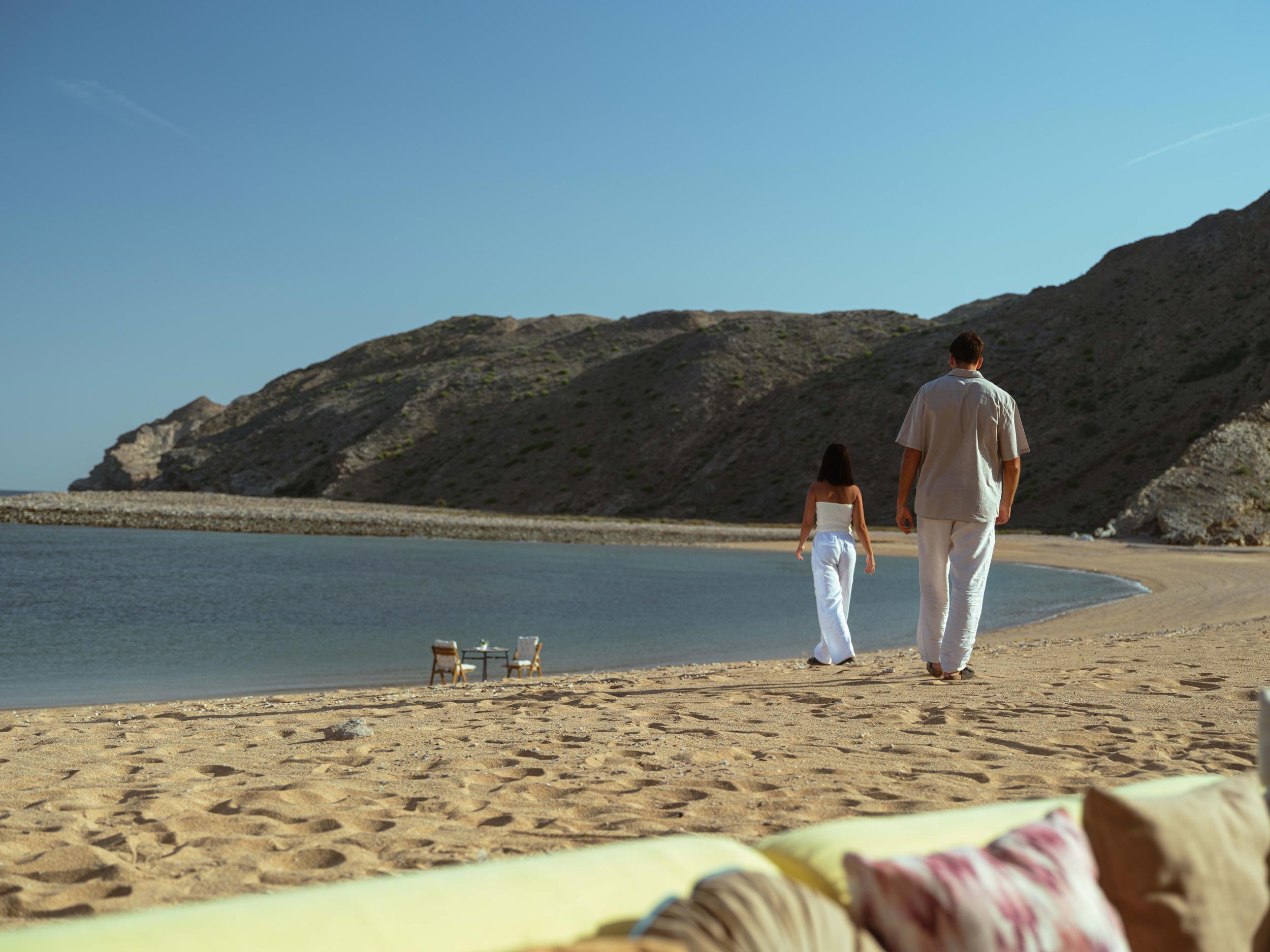 A man and woman walking along a beach, with chairs and a table set up in the distance and hills in the background.