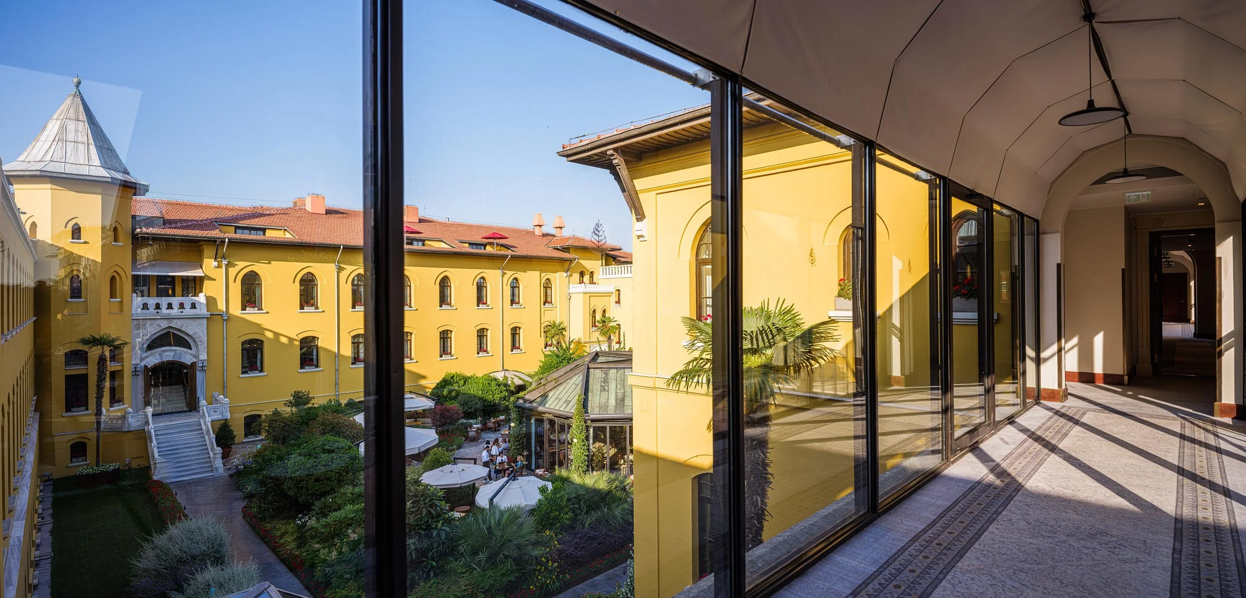 View of a yellow building and outdoor garden seen through large glass windows from an indoor corridor with decorative tiled flooring.