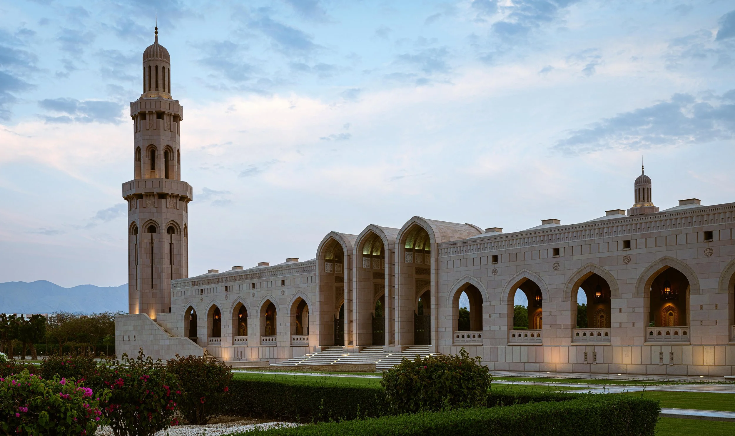 A large, ornate mosque with a tall minaret, multiple arches, and intricate architectural details, set against a backdrop of mountains and a cloudy sky.