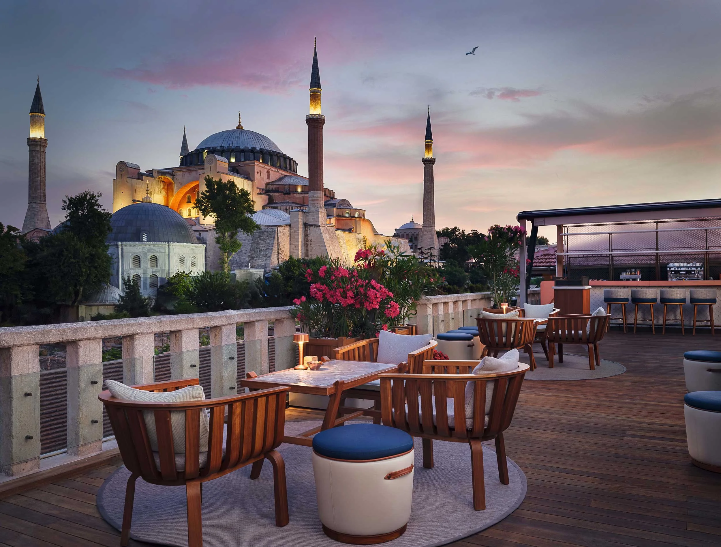Rooftop restaurant terrace of Four Seasons Istanbul Hotel at Sultanahmet with wooden chairs, tables, and pink flowers, overlooking the historic Hagia Sophia mosque illuminated at sunset with a bird flying in the sky.