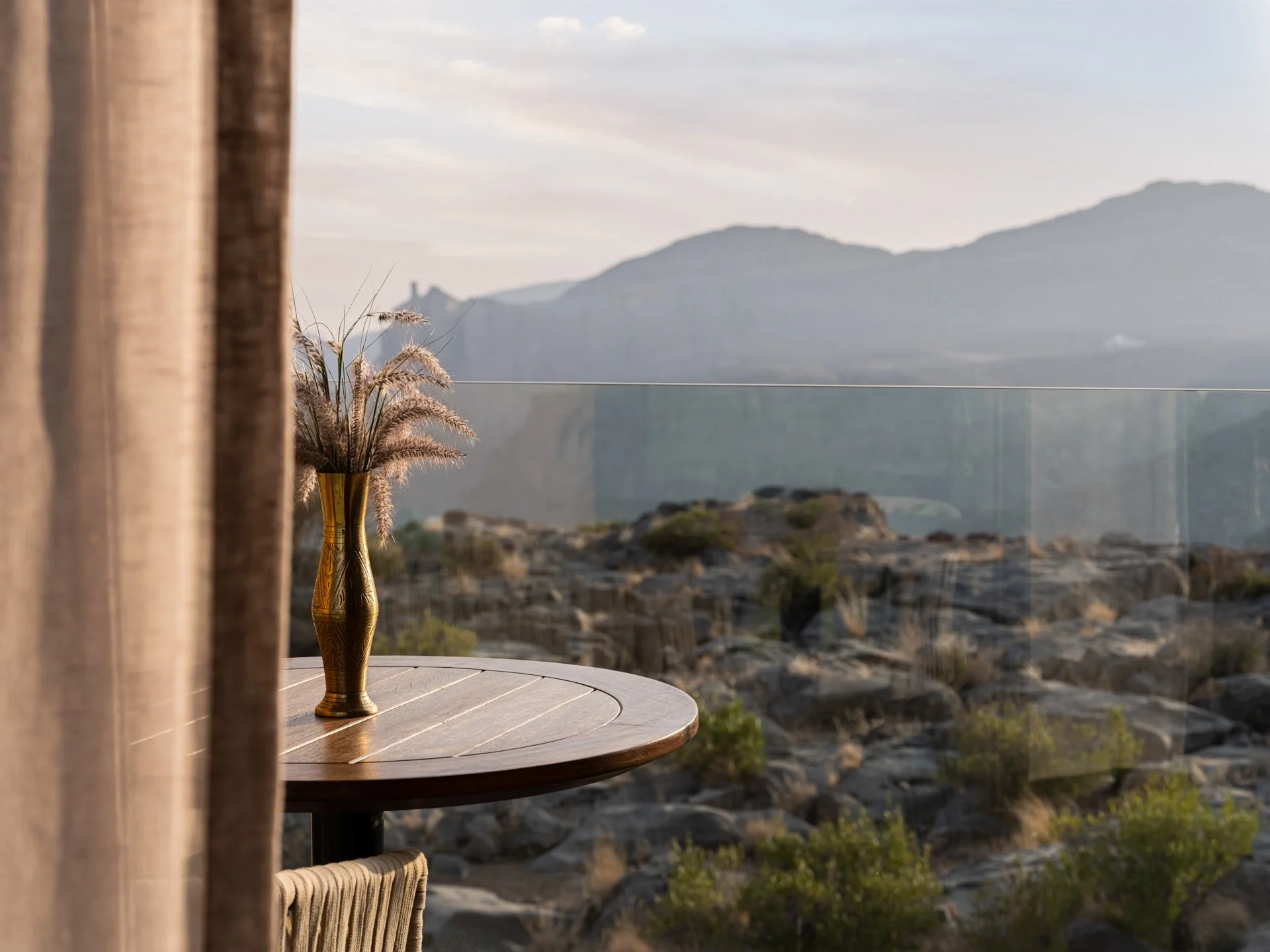 Vase with dried grasses on a wooden table overlooking a rocky landscape and mountains.