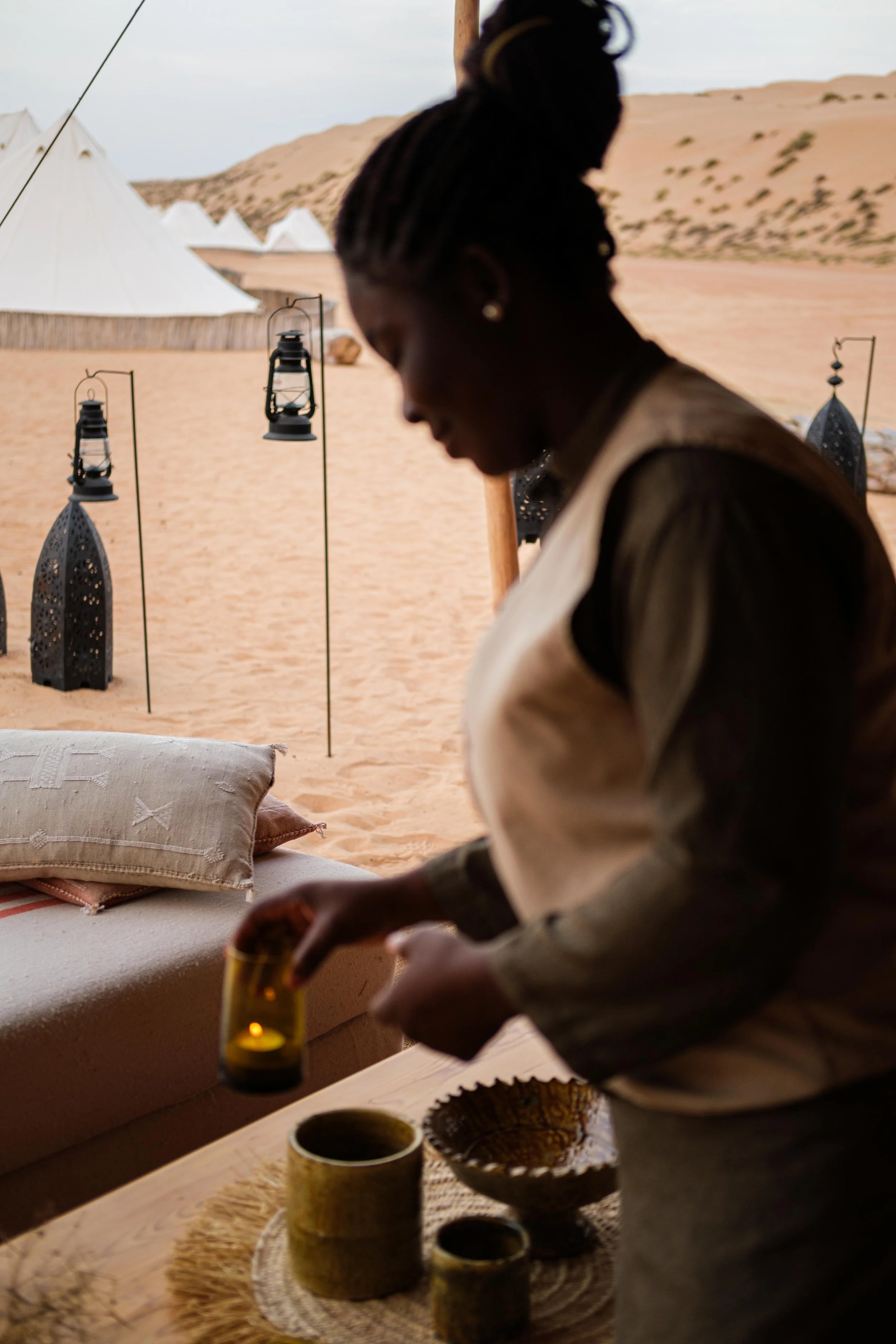 Person arranging items on a wooden table in a desert camp setting with tents and lanterns.