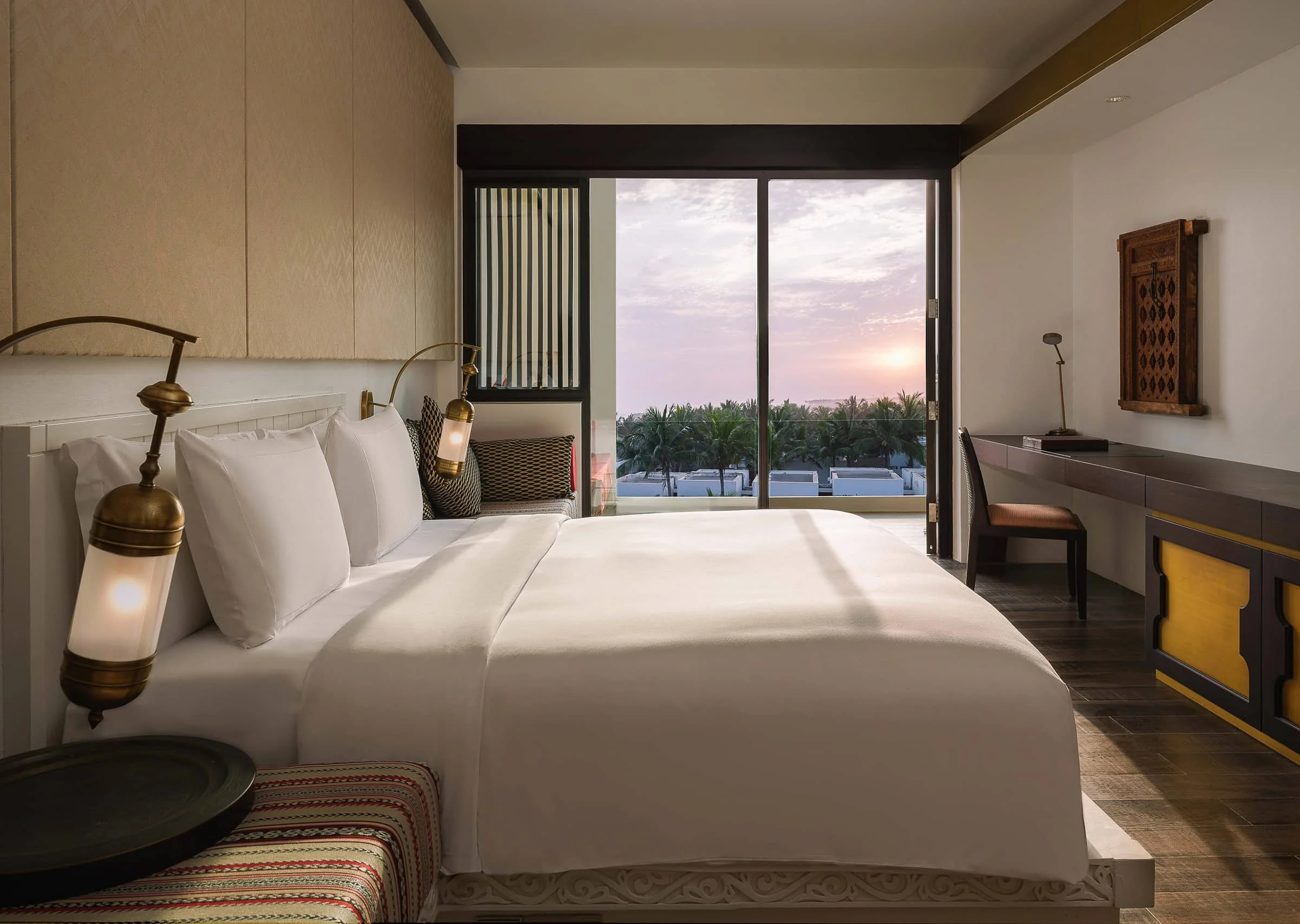 A hotel room with a large bed, bedside lamps, and a view of palm trees and a sunset through sliding glass doors.