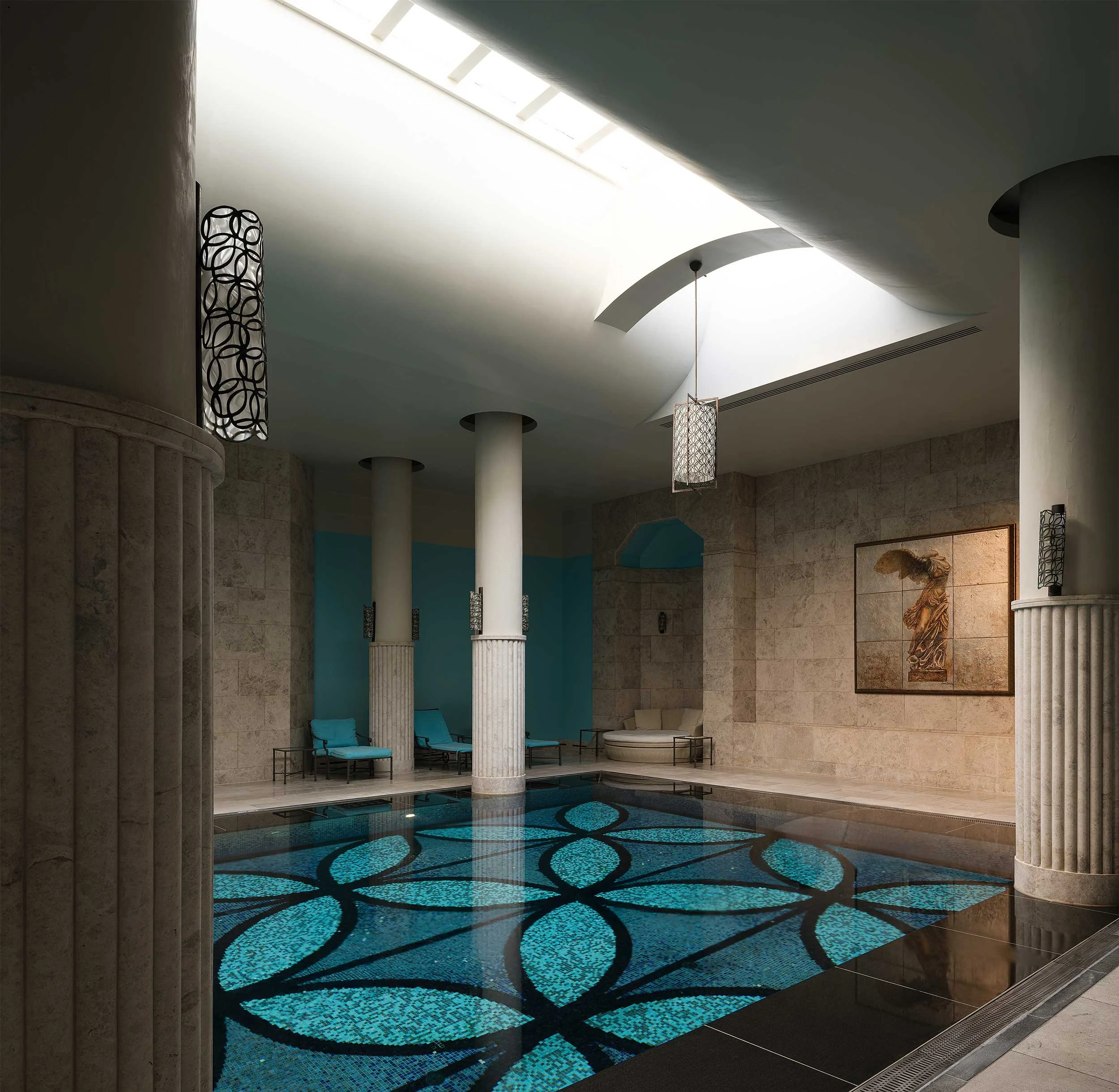 Indoor swimming pool area with blue seating, stone walls, columns, and artwork on the wall.