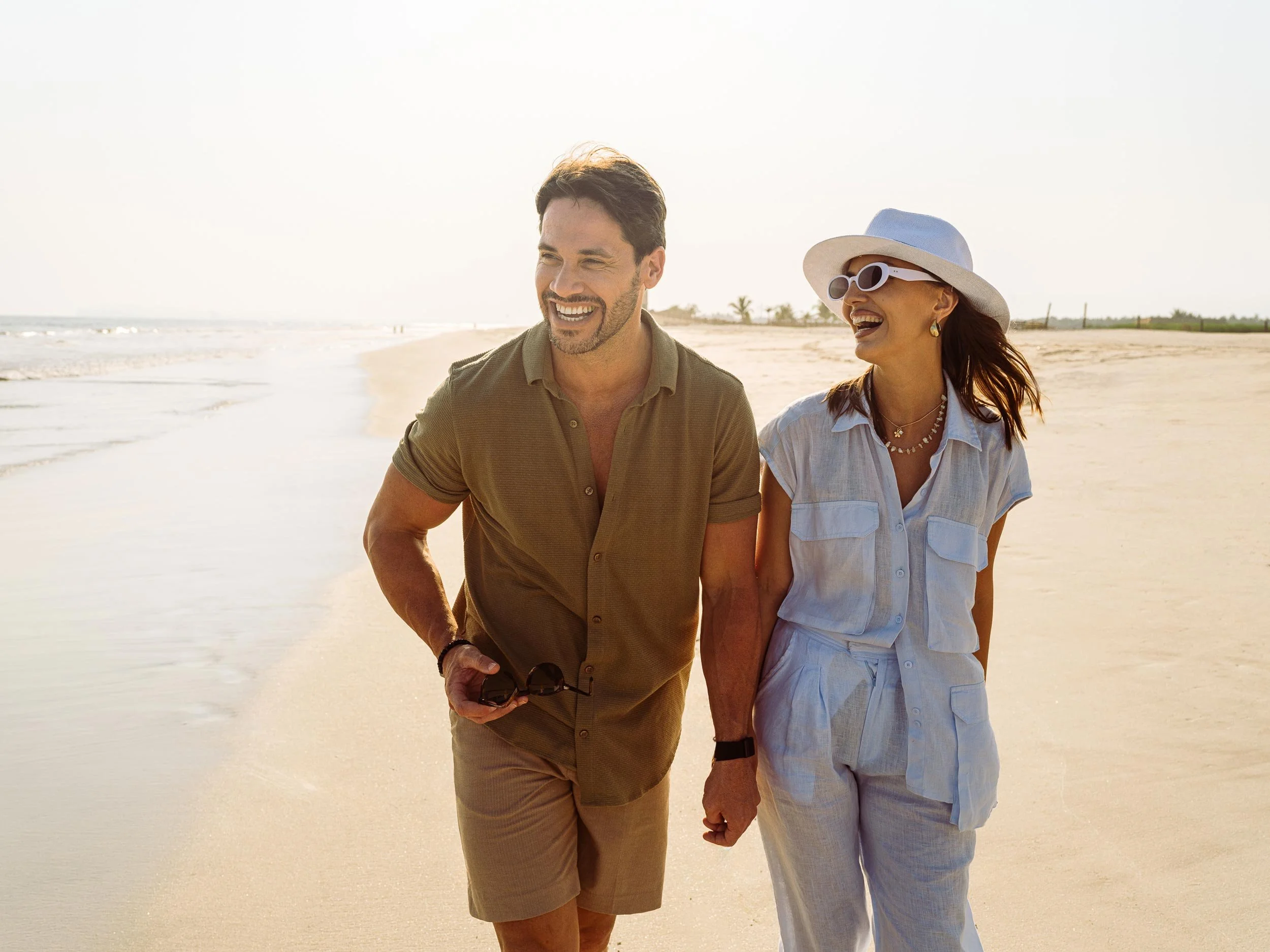A smiling man and woman walking on a beach holding hands, enjoying sunny weather with the ocean in the background.