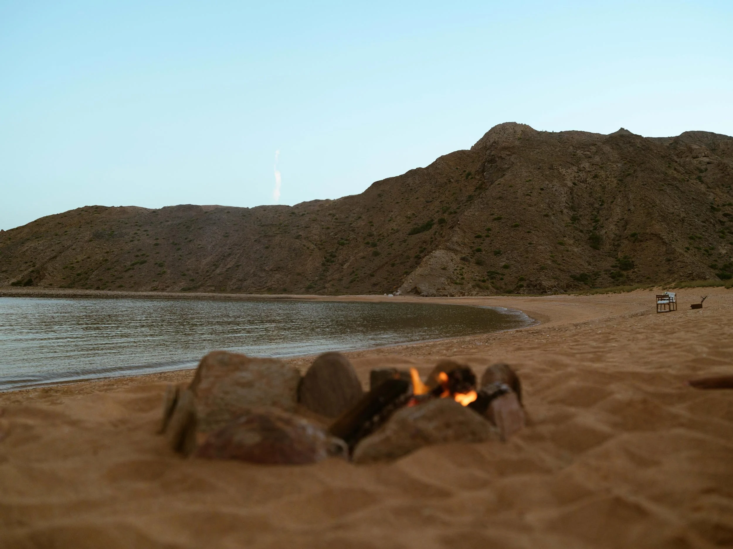 A peaceful beach scene with a small campfire in the foreground, sandy shore, calm water, mountainous terrain in the background, and a clear sky.