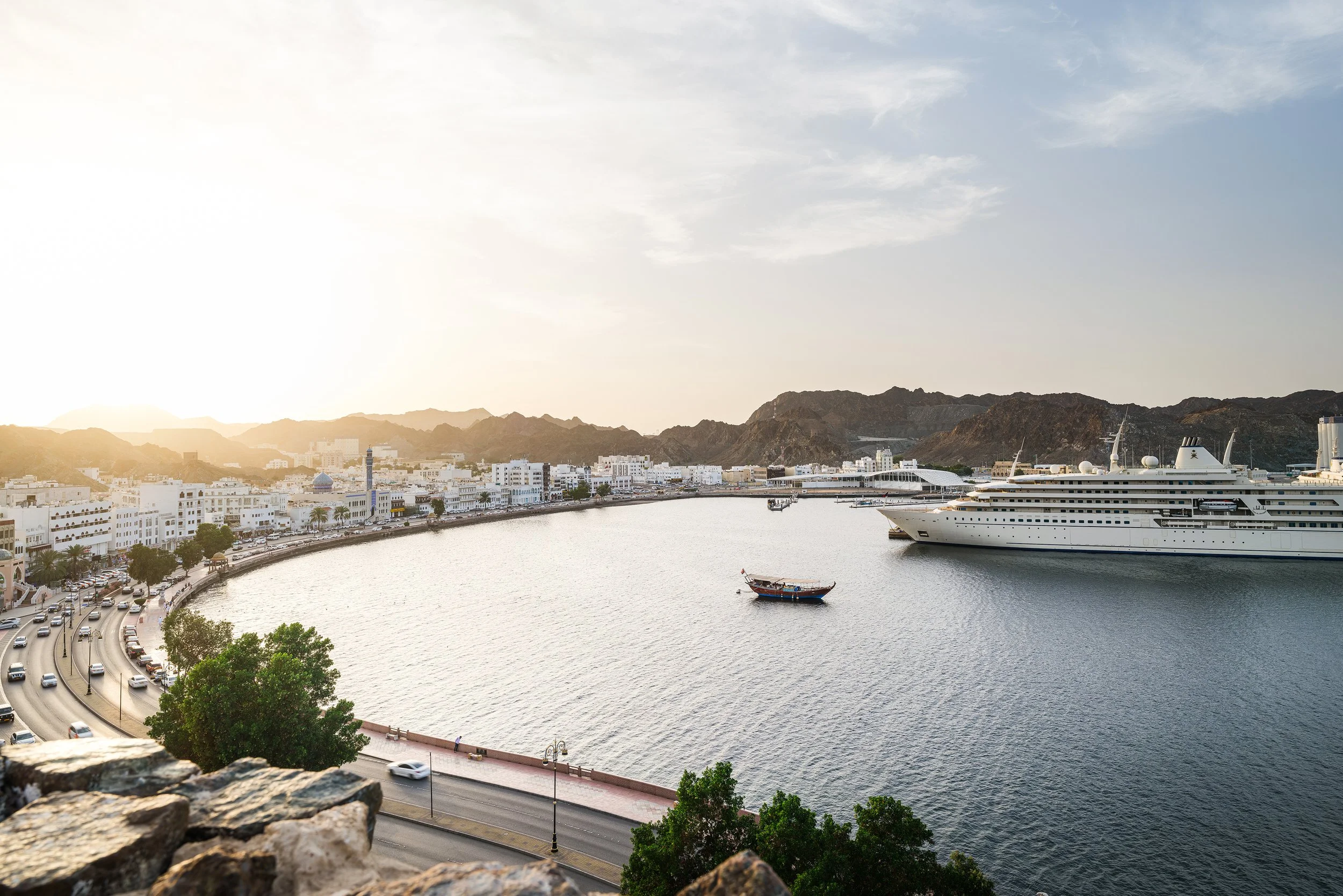 A harbor with a large cruise ship docked near the city. Smaller boats float in the water. The city has white buildings, mountains in the background, and a road with cars along the waterfront. The sky is partly cloudy with sunlight.