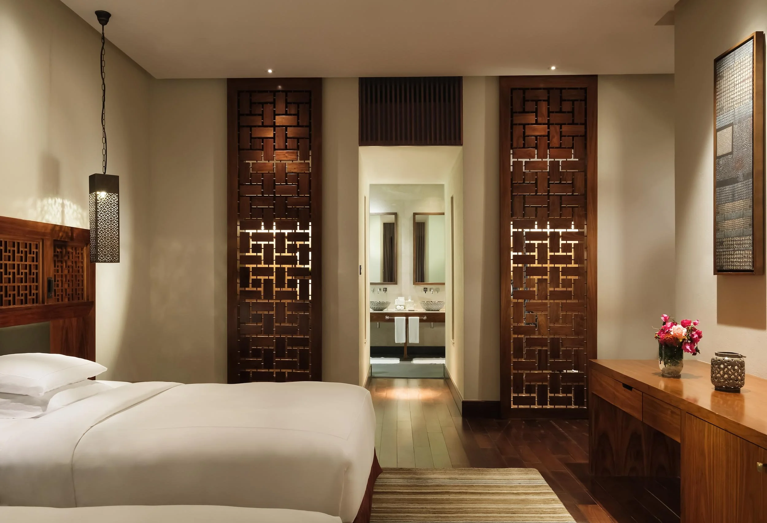 A hotel room with a king-sized bed, wooden accents, decorative wooden screens, a side table with a flower arrangement, and an open doorway leading to a bathroom with two sinks and mirrors.
