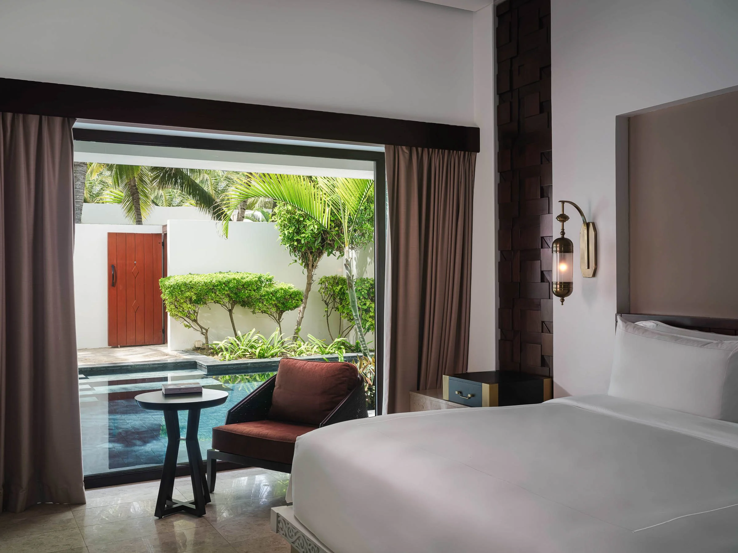 Modern hotel room with a large bed, nightstand, and wall-mounted lamp, opening to a private outdoor patio with a pool, plants, and a red door.