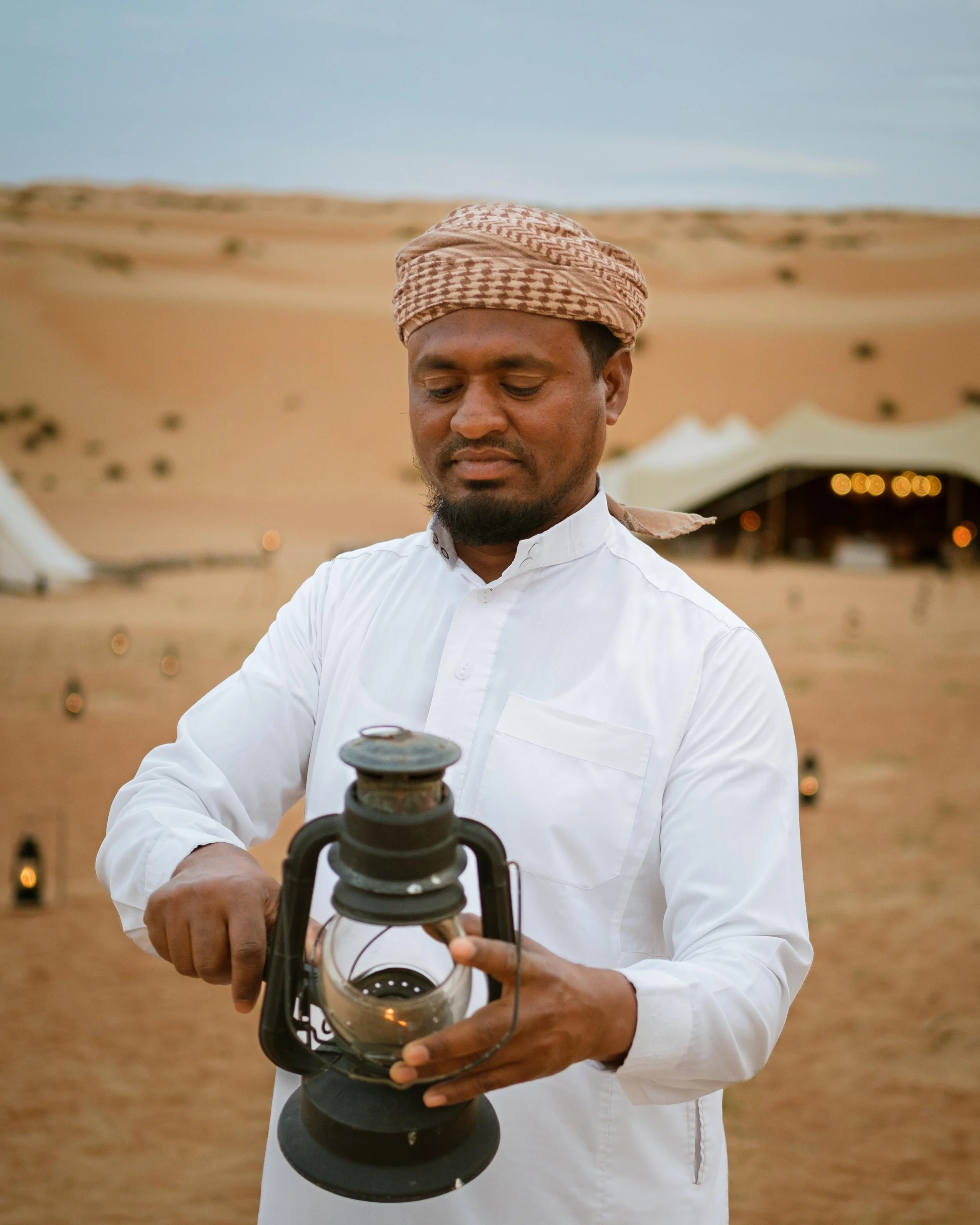 A man in traditional attire lighting a lantern in a desert setting with tents in the background.
