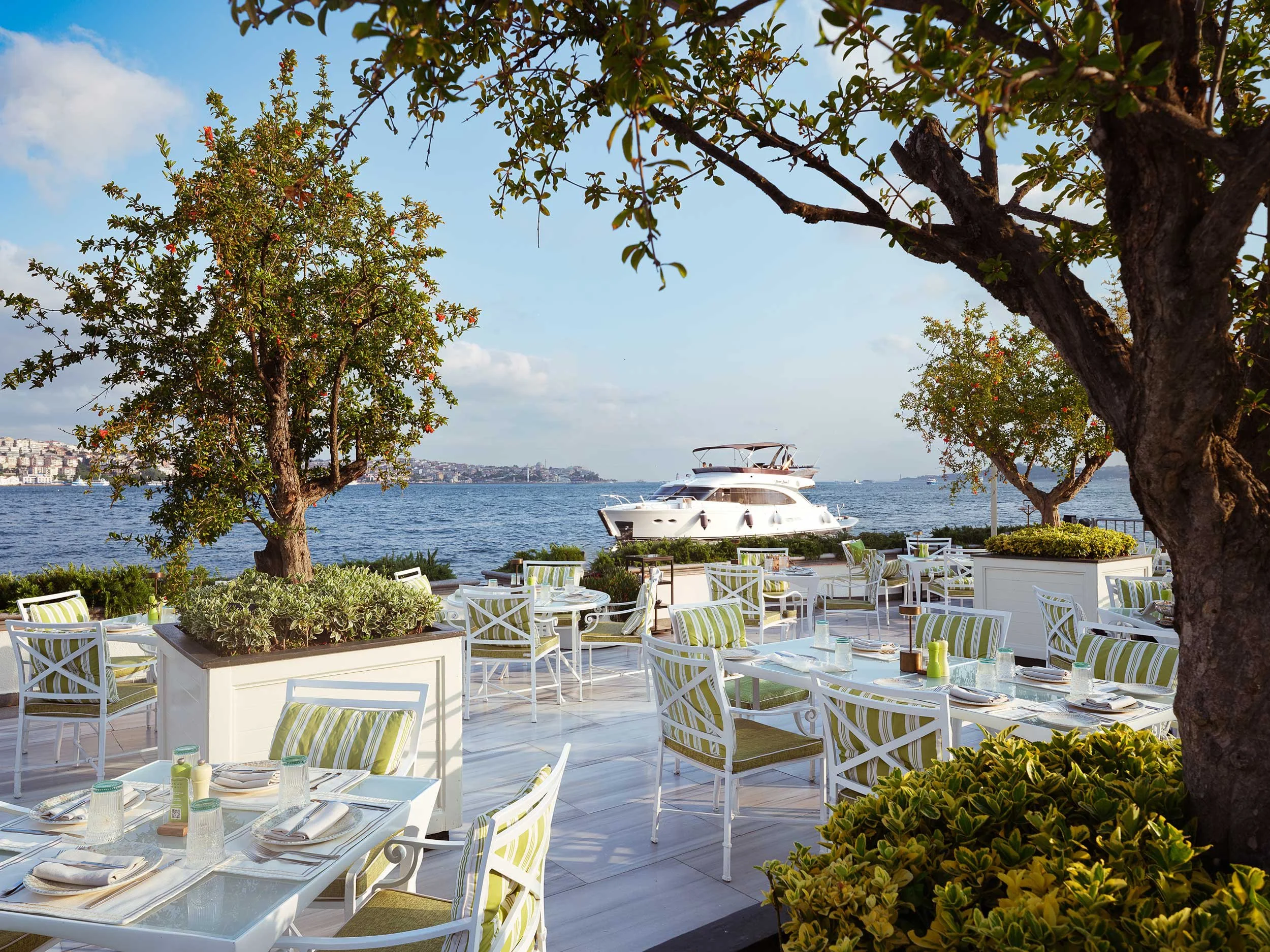 Outdoor waterfront dining area with white tables and chairs, surrounded by trees and greenery, overlooking a body of water with a yacht in the background.
