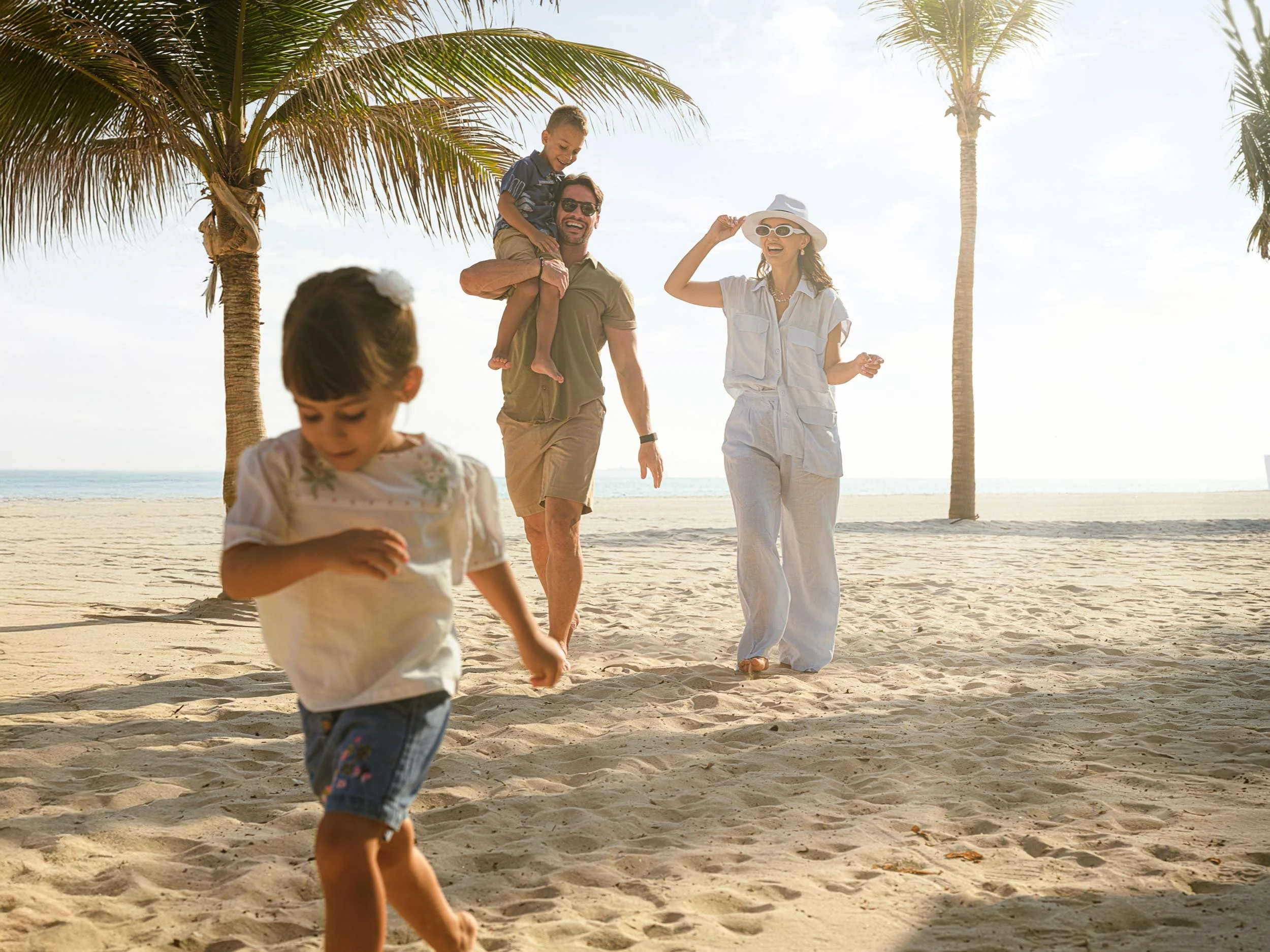 A family enjoying a day at the beach with palm trees, two children, a man, and a woman walking and playing in the sand under a sunny sky.