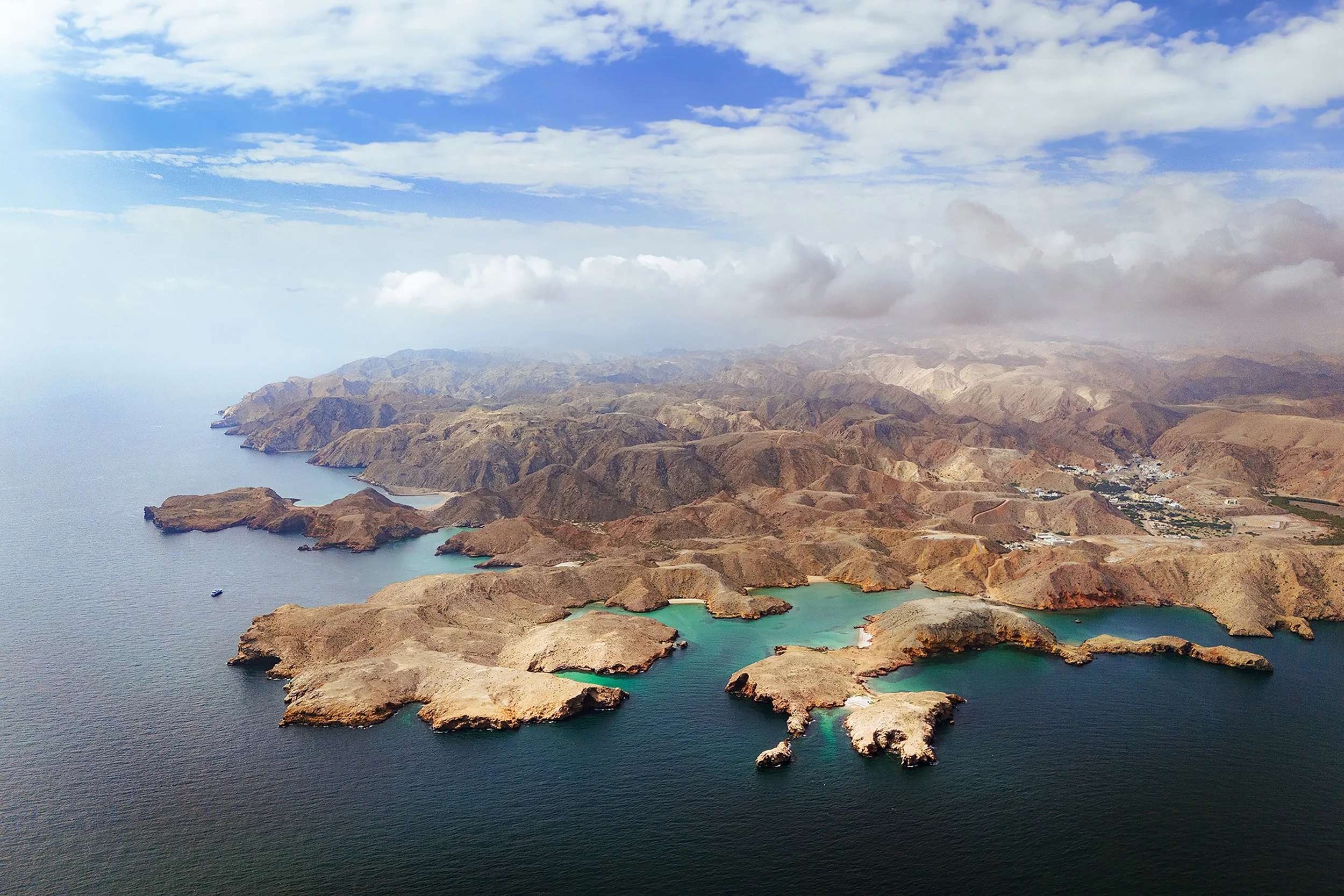 Aerial view of a rugged coastline with rocky cliffs and coves along a blue ocean, surrounded by hilly, arid terrain under a partly cloudy sky.