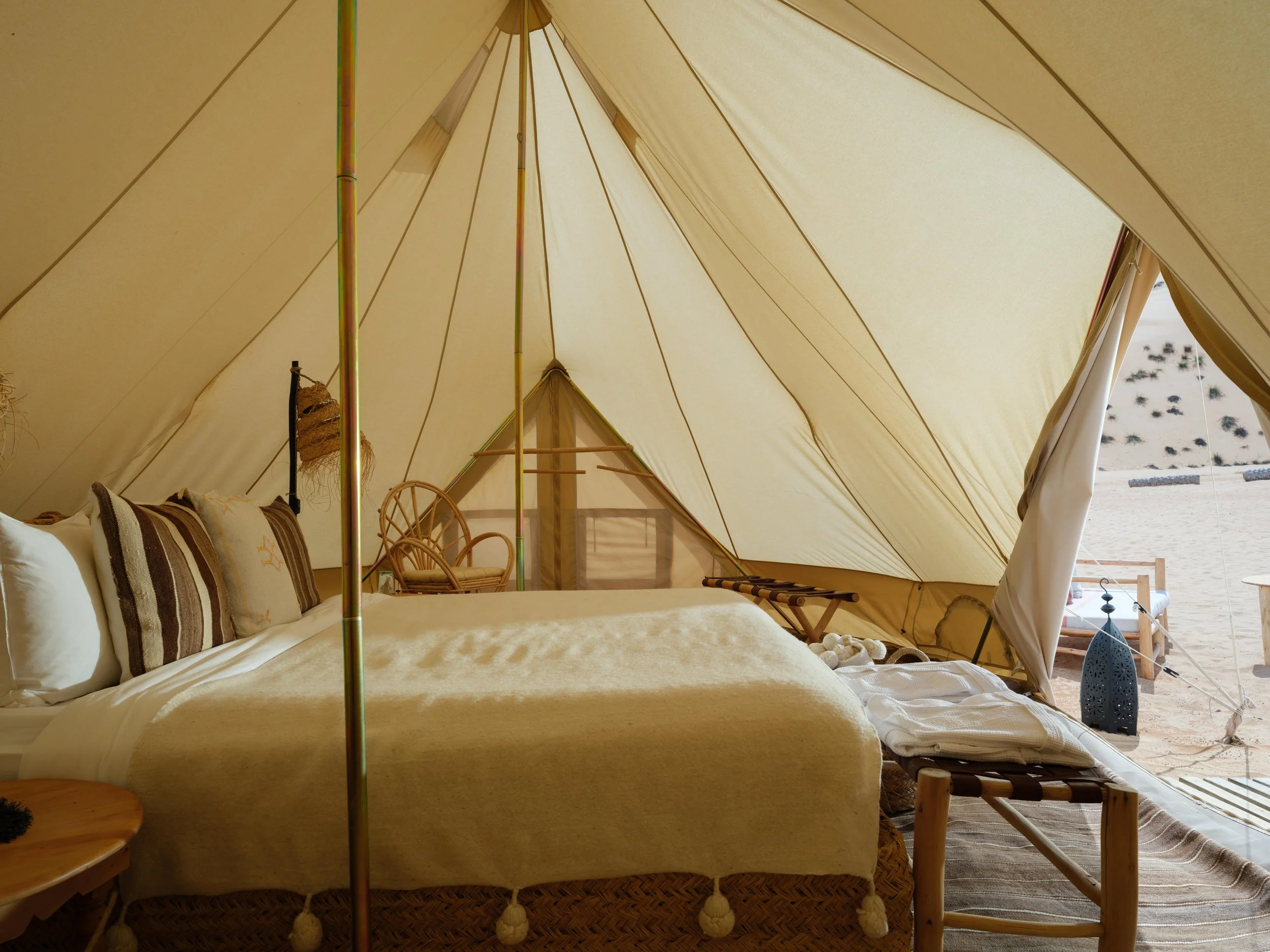 Luxurious interior of a glamping tent with a bed, woven chairs, and blankets, set in a desert landscape.
