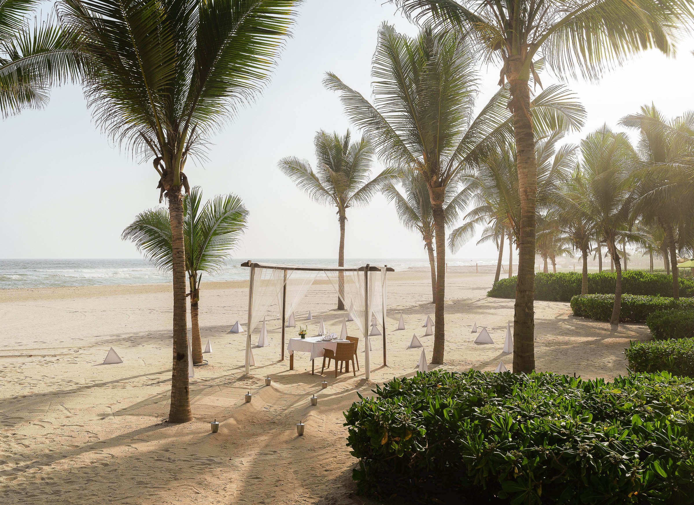 A wedding setup on a sandy beach with palm trees, a small canopy, a table with chairs, and decorative lanterns, overlooking the ocean.