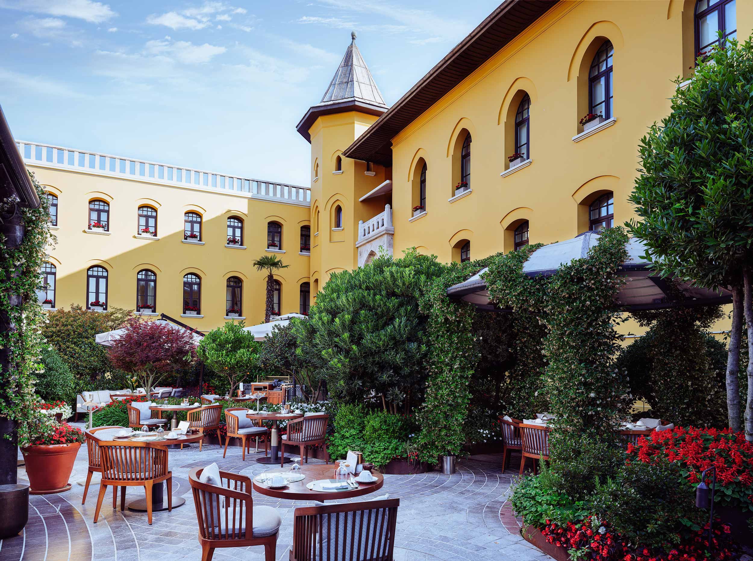 Outdoor patio with dining tables and chairs, surrounded by lush green plants and trees, with a yellow building with arched windows in the background.