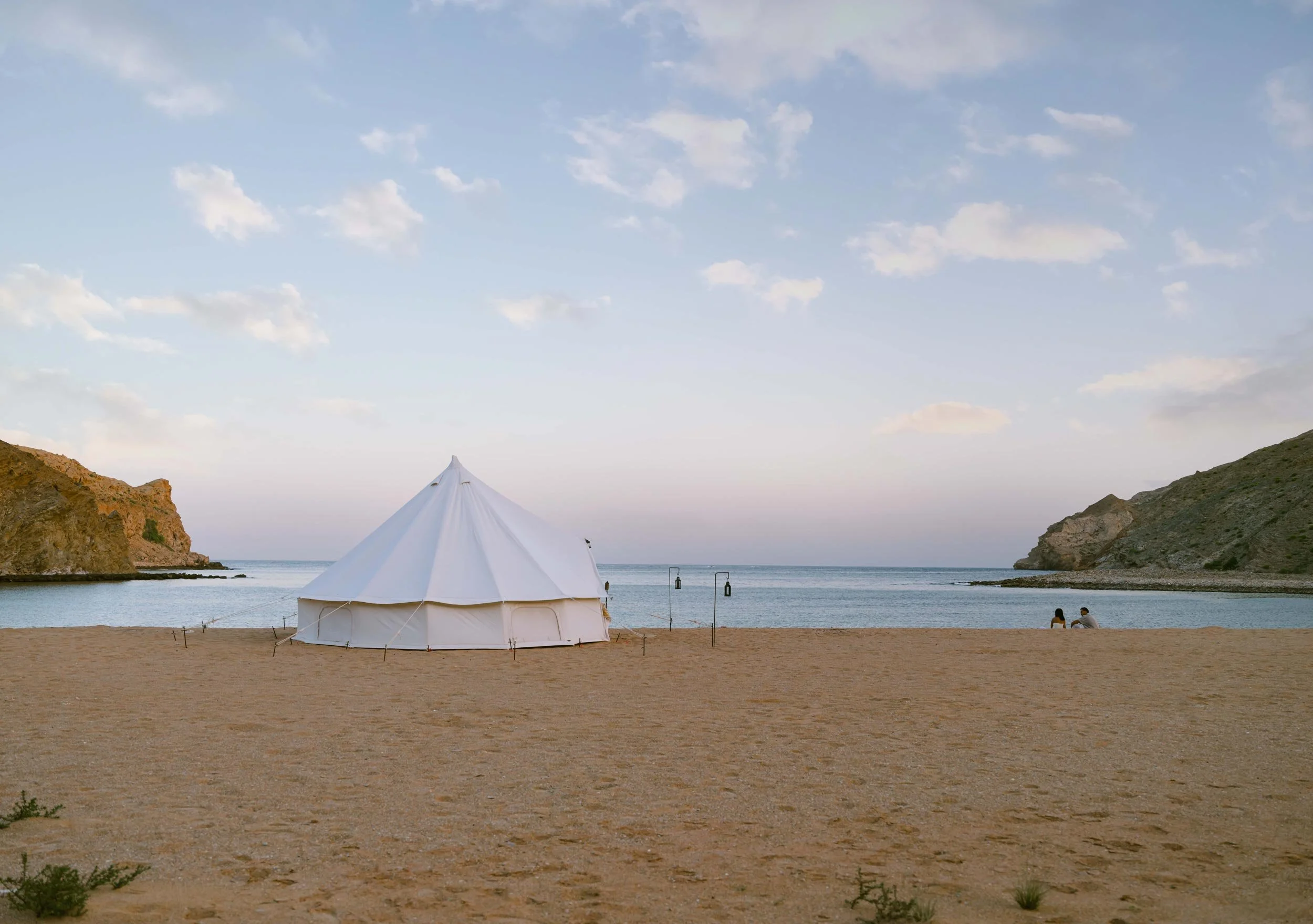 A white tent set up on a sandy beach near the water, flanked by rocky hills on either side, with two people sitting near the shoreline and a few lampposts standing in the sand in Bandar al Khiran - Oman