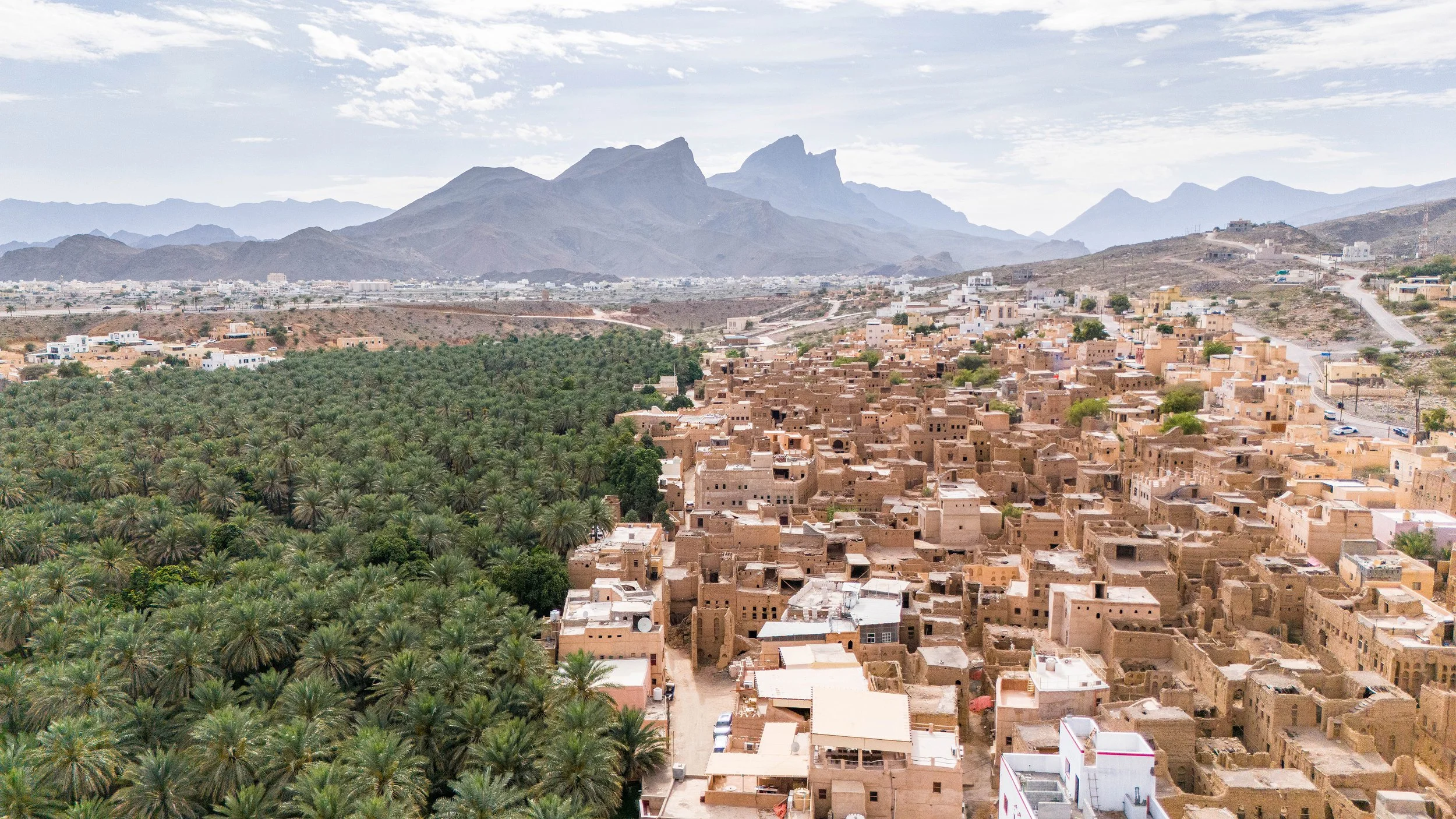 Aerial view of a desert town with beige buildings on the right and a green palm grove on the left, with mountains in the distance under a partly cloudy sky.