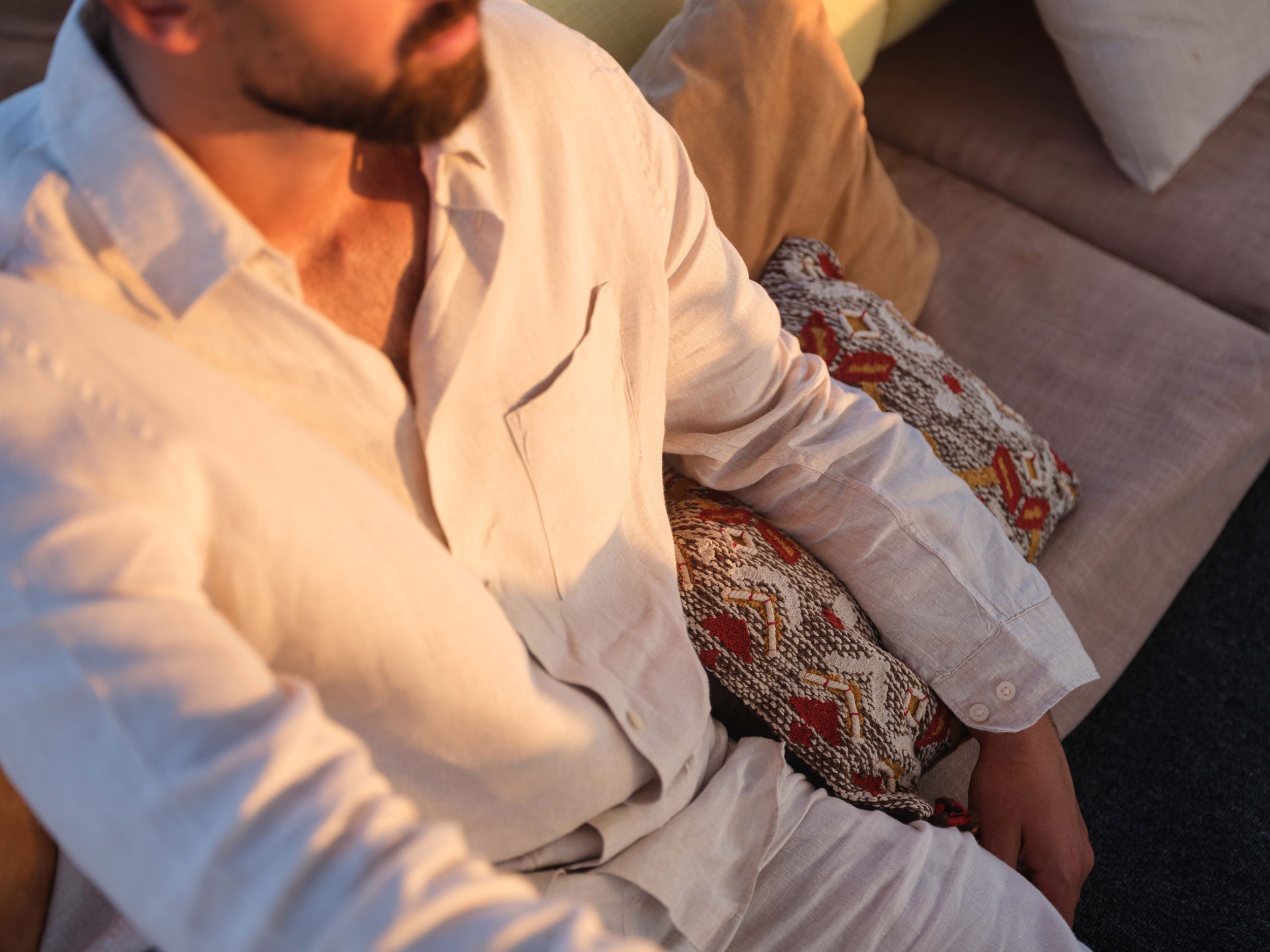 A man wearing a light-colored shirt sitting on a beige couch with patterned pillows.