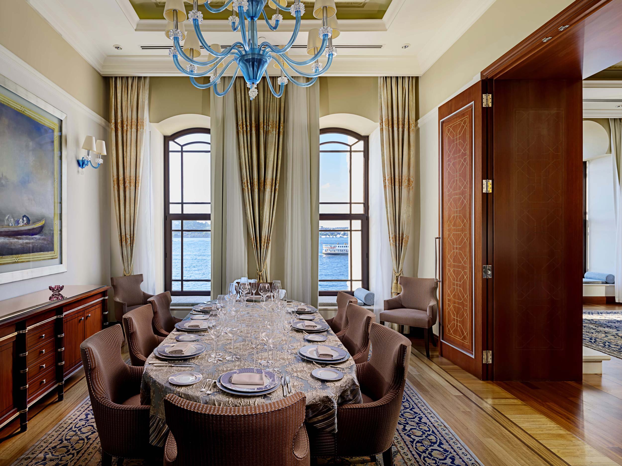 Elegant dining room with a large table set for a meal, overlooking water through tall windows, with chandelier and curtains.