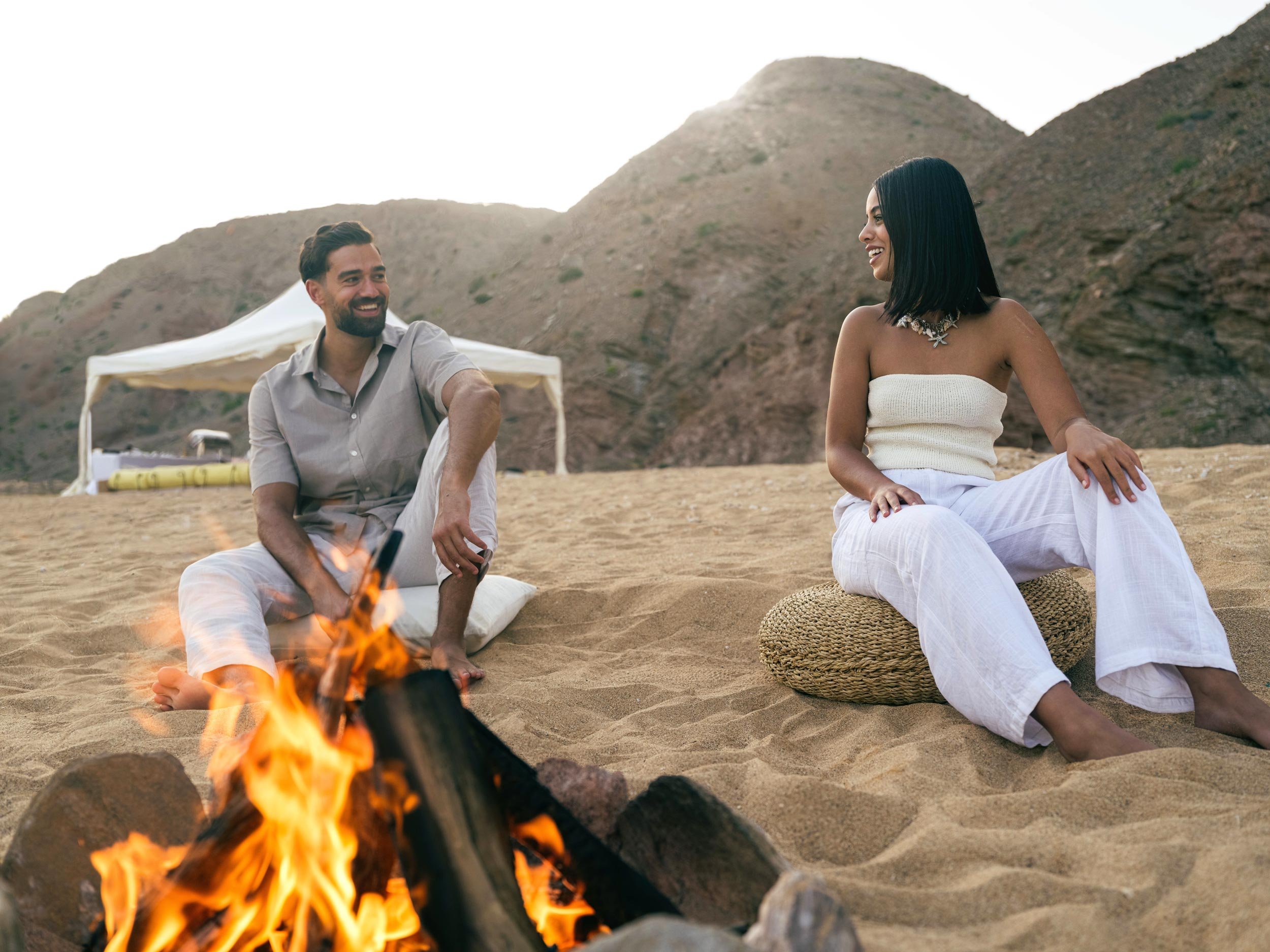 A man and woman sitting around a campfire on a sandy bandar al khiran beach with hills in the background, smiling and enjoying each other's company.