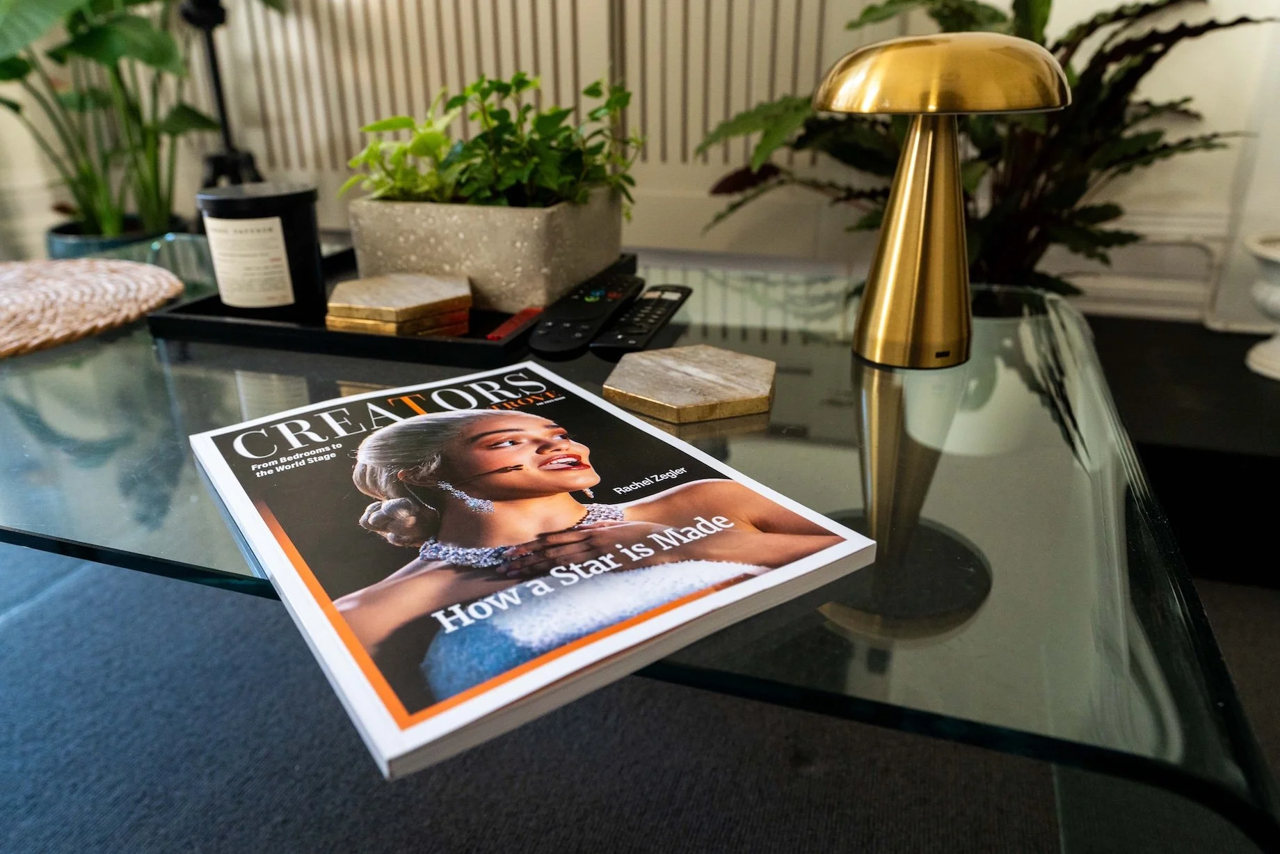 A magazine titled "Creatrors Trove" featuring a man with a pink bandana holding his finger to his lips, pointing to the magazine. In the foreground, a blue bubble water bottle and part of a camera are visible on a wooden table against a pink background.