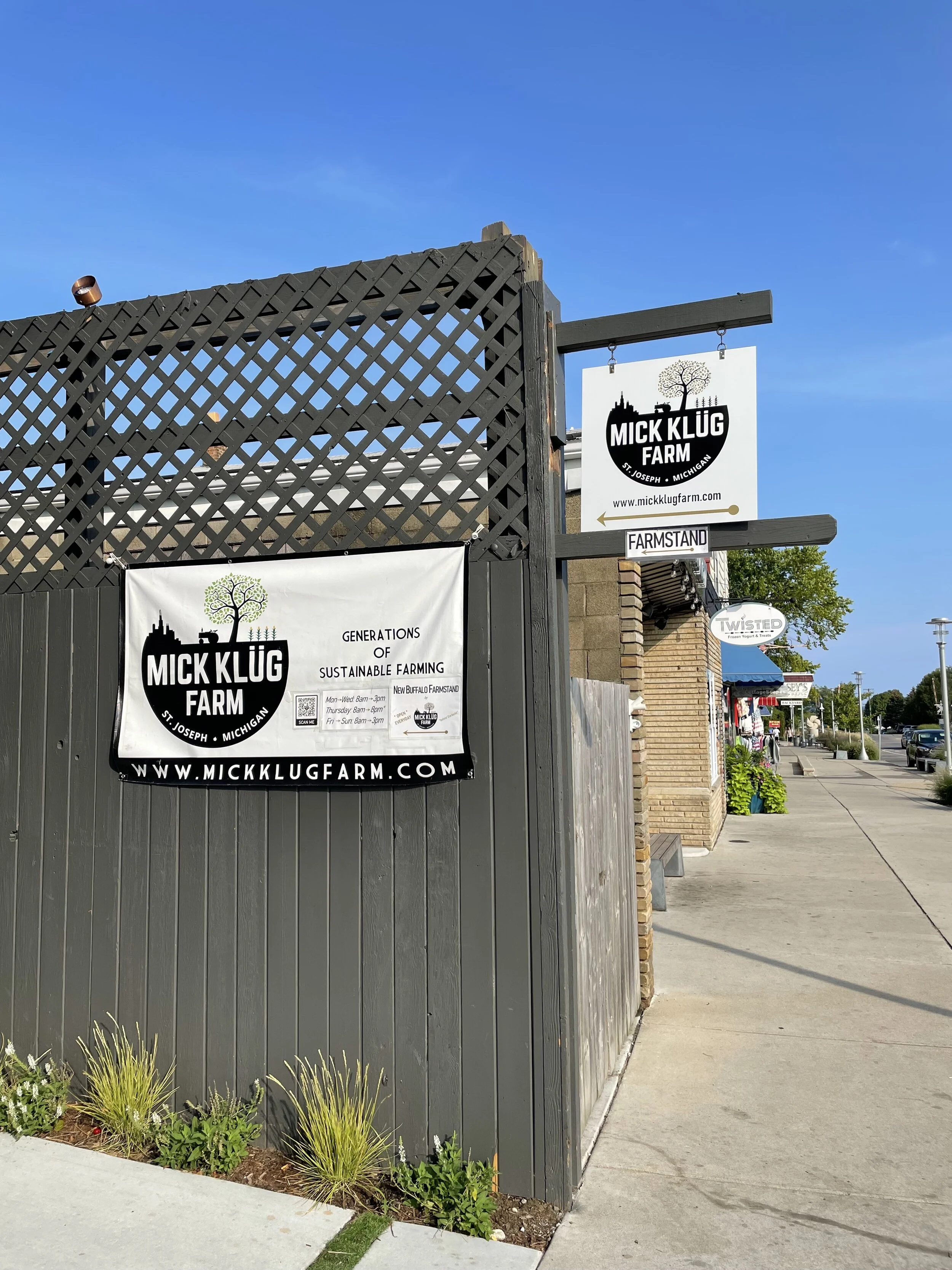 Sign for Mick Klug Farm on a street corner in St. Joseph, Michigan. The sign is attached to a gray wooden fence with surrounding small plants and flowers.