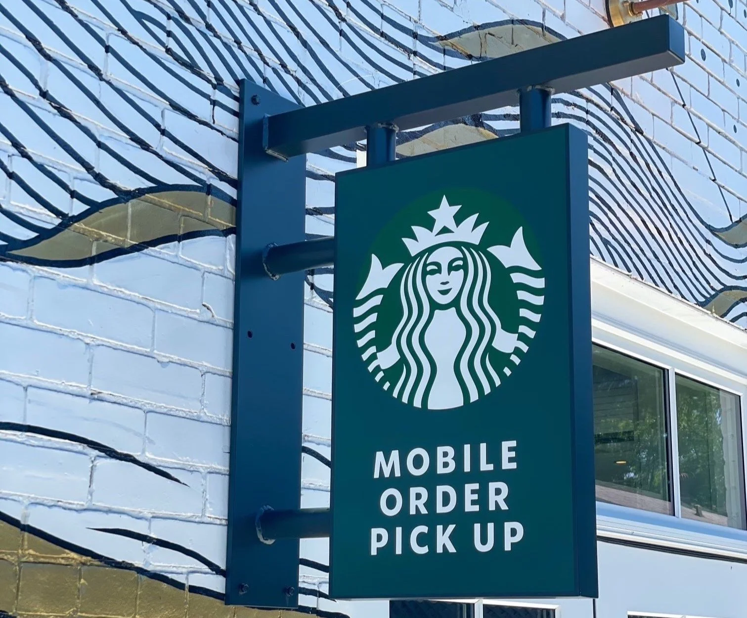 Starbucks sign indicating mobile order pickup outside a building with a white and gold brick wall and colorful mural