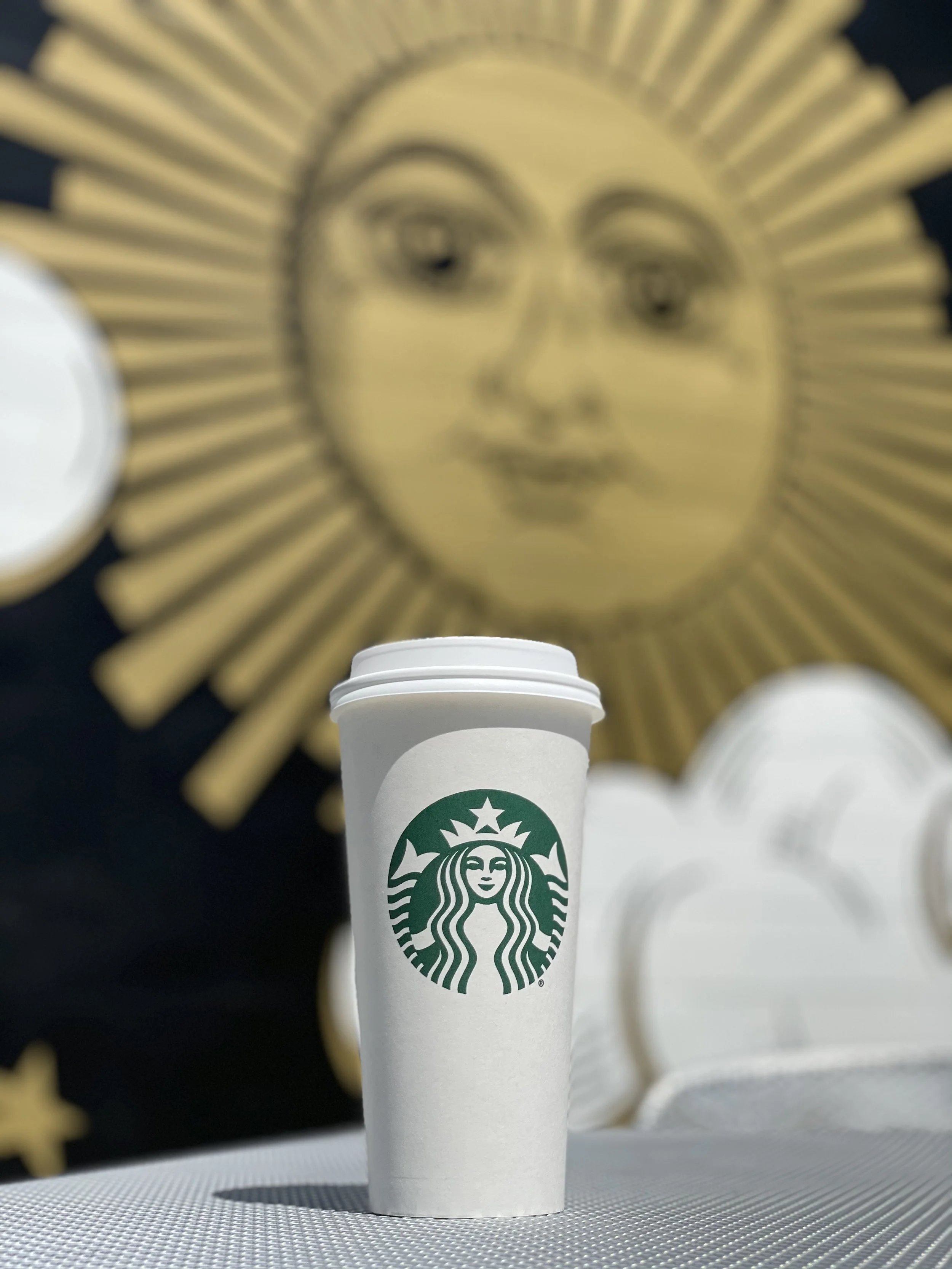 Starbucks disposable coffee cup with a green logo on a white background, in front of a large decorative sun face with a metallic gold finish and radiating rays.