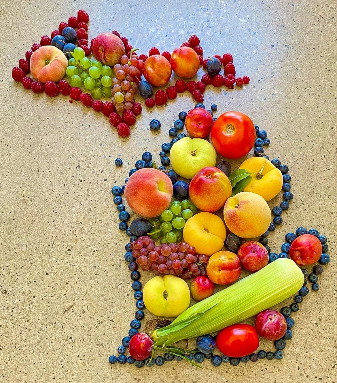 Arrangement of various fresh fruits including peaches, apples, grapes, blueberries, tomatoes, and a cobs of corn on a beige speckled surface, forming the shape of the number five.