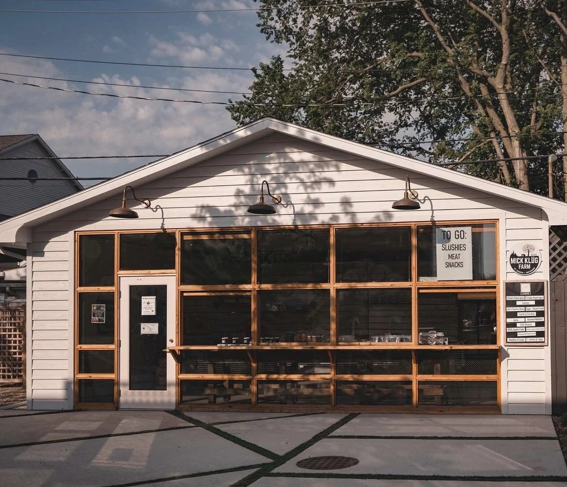 Small white building with large front windows and a sign that reads 'TO GO: SLUSHIES, MEAT, SNACKS', with a wooden frame around the windows and door, two exterior wall lamps, and a tree casting shadows on the building.