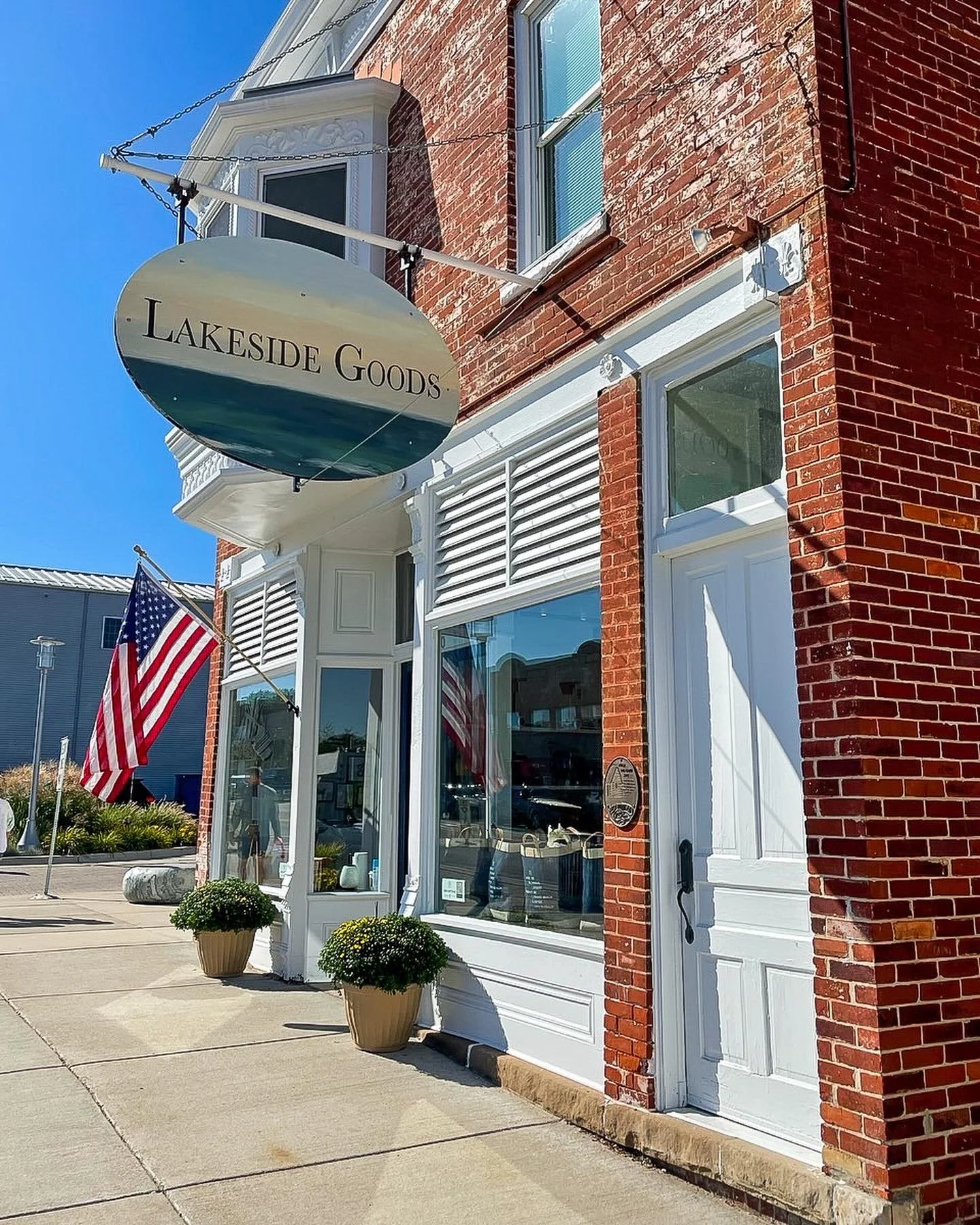The exterior of a brick store called Lakeside Goods with a hanging sign, American flags, and potted plants outside, under a clear blue sky.